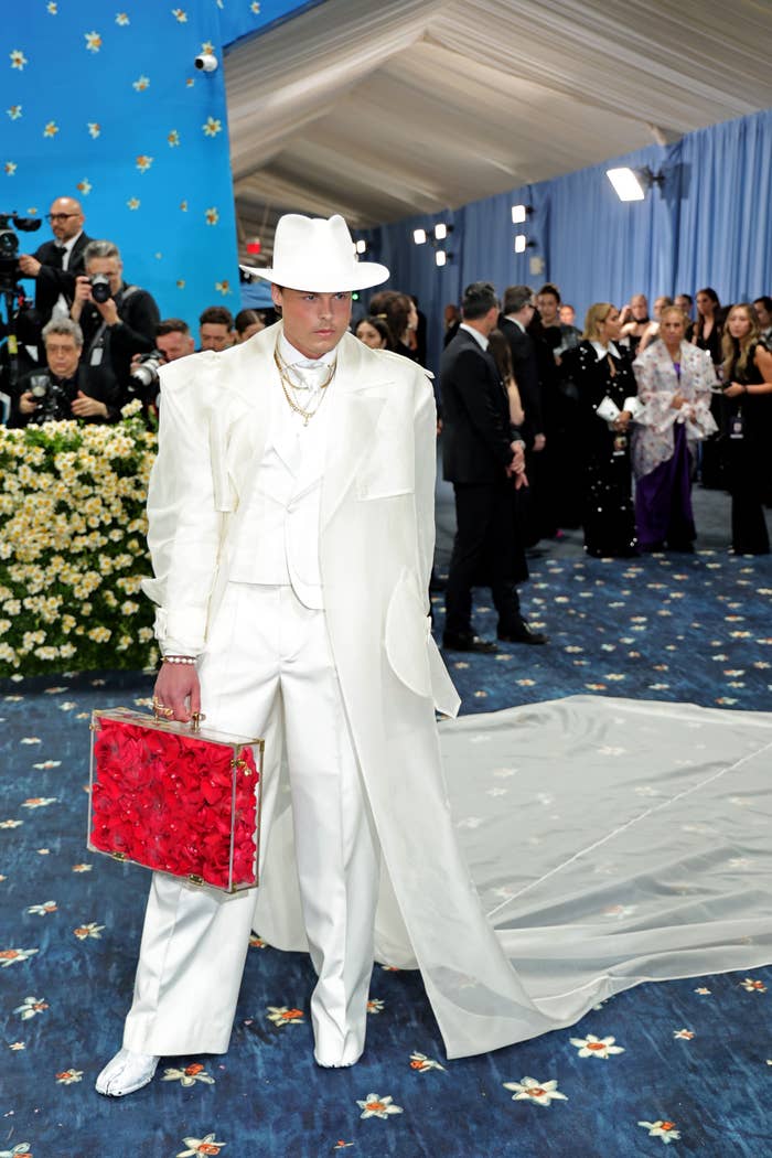 Person wearing a achromatic suit with a agelong bid and achromatic hat, holding a reddish floral container connected a bluish carpet astatine a personage event