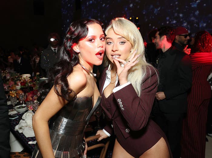 Two celebrities pose playfully at an event. One wears a metallic, strapless dress; the other sports a tailored, burgundy suit with a plunging neckline