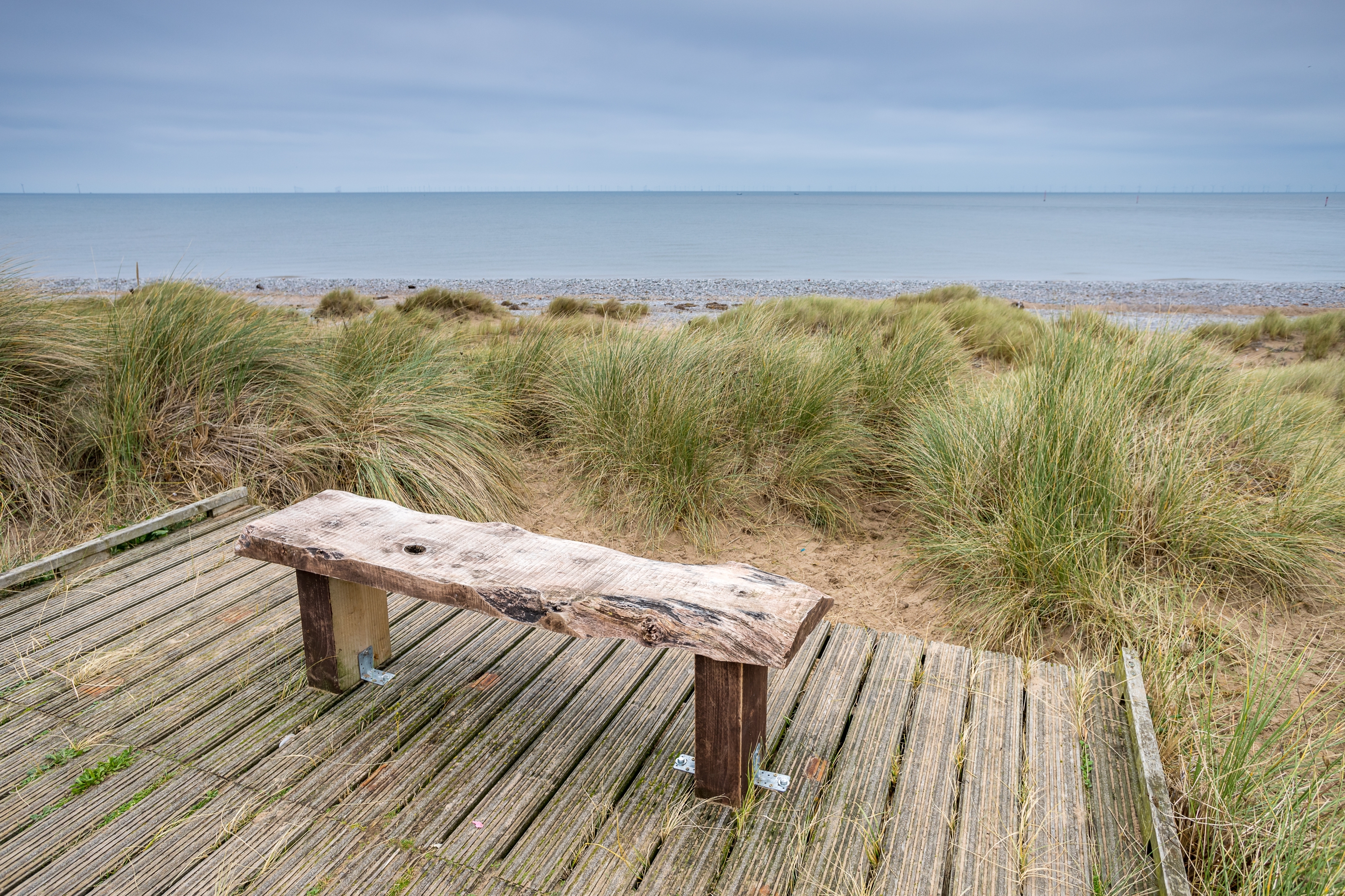 A weathered wooden bench on a boardwalk overlooks a grassy dune and calm sea under a cloudy sky