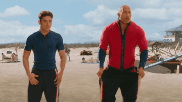 Two men in form-fitting athletic gear stand on a beach, appearing to discuss something