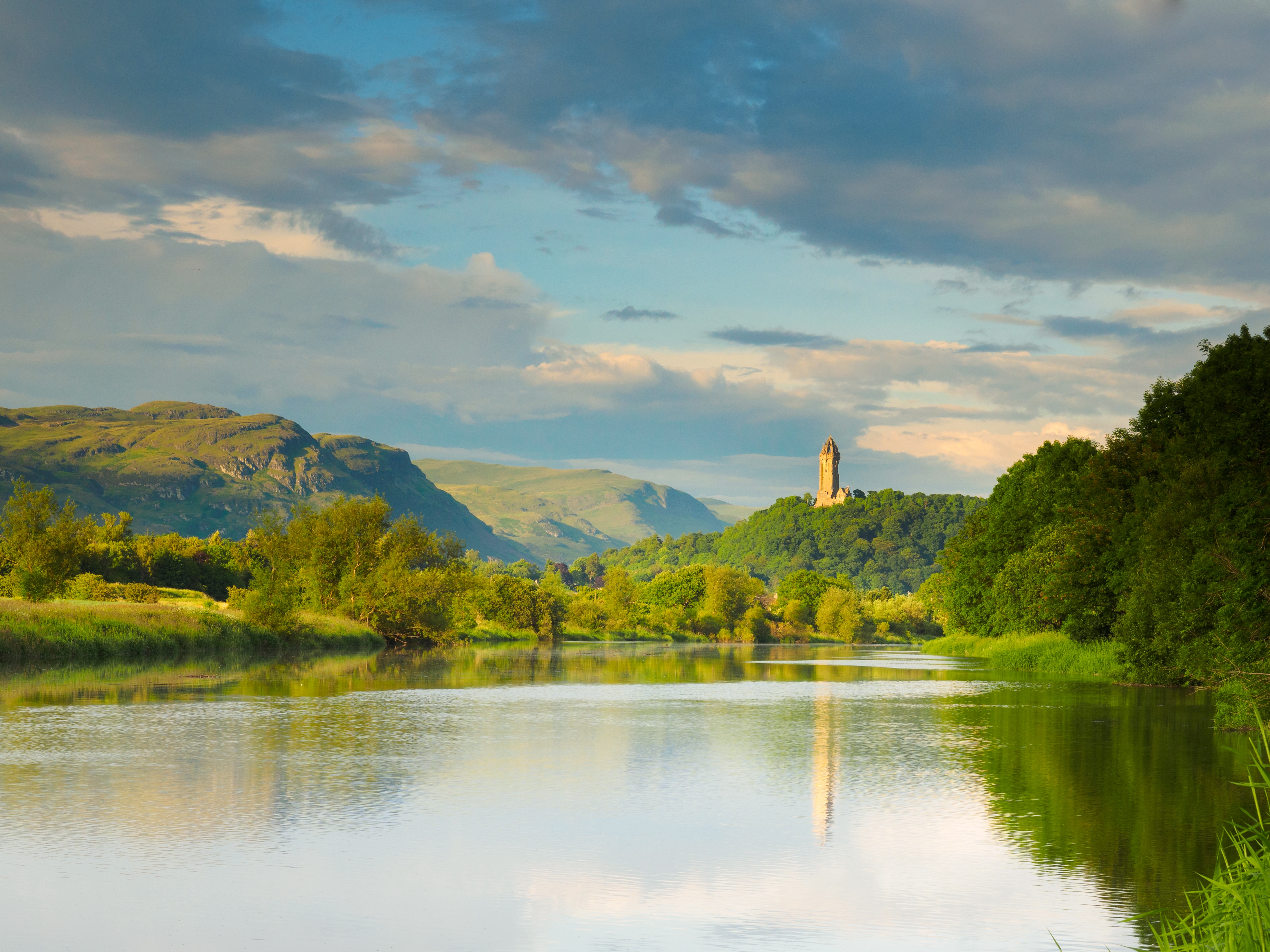 A tranquil river scene with a historic tower on a distant hill, surrounded by lush greenery and hills under a partly cloudy sky
