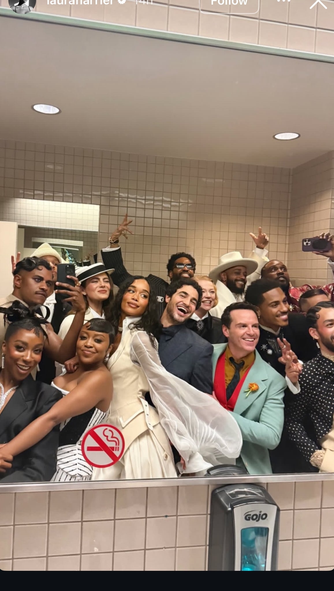 Group of people in a bathroom selfie at an event, dressed in formal and stylish attire, some wearing hats and accessories, smiling and posing