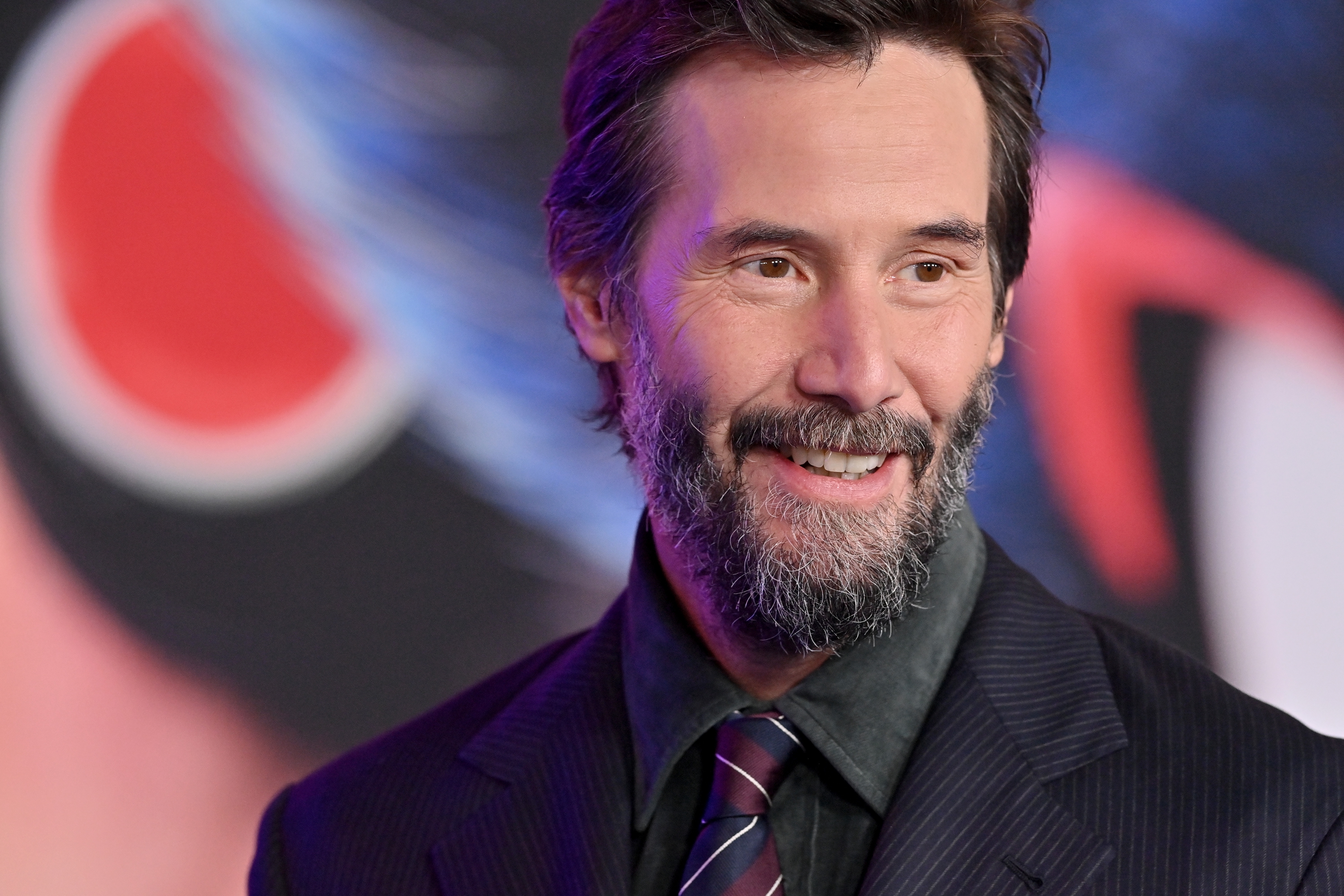 Keanu Reeves with a beard smiles while wearing a dark suit with a striped tie at a formal event
