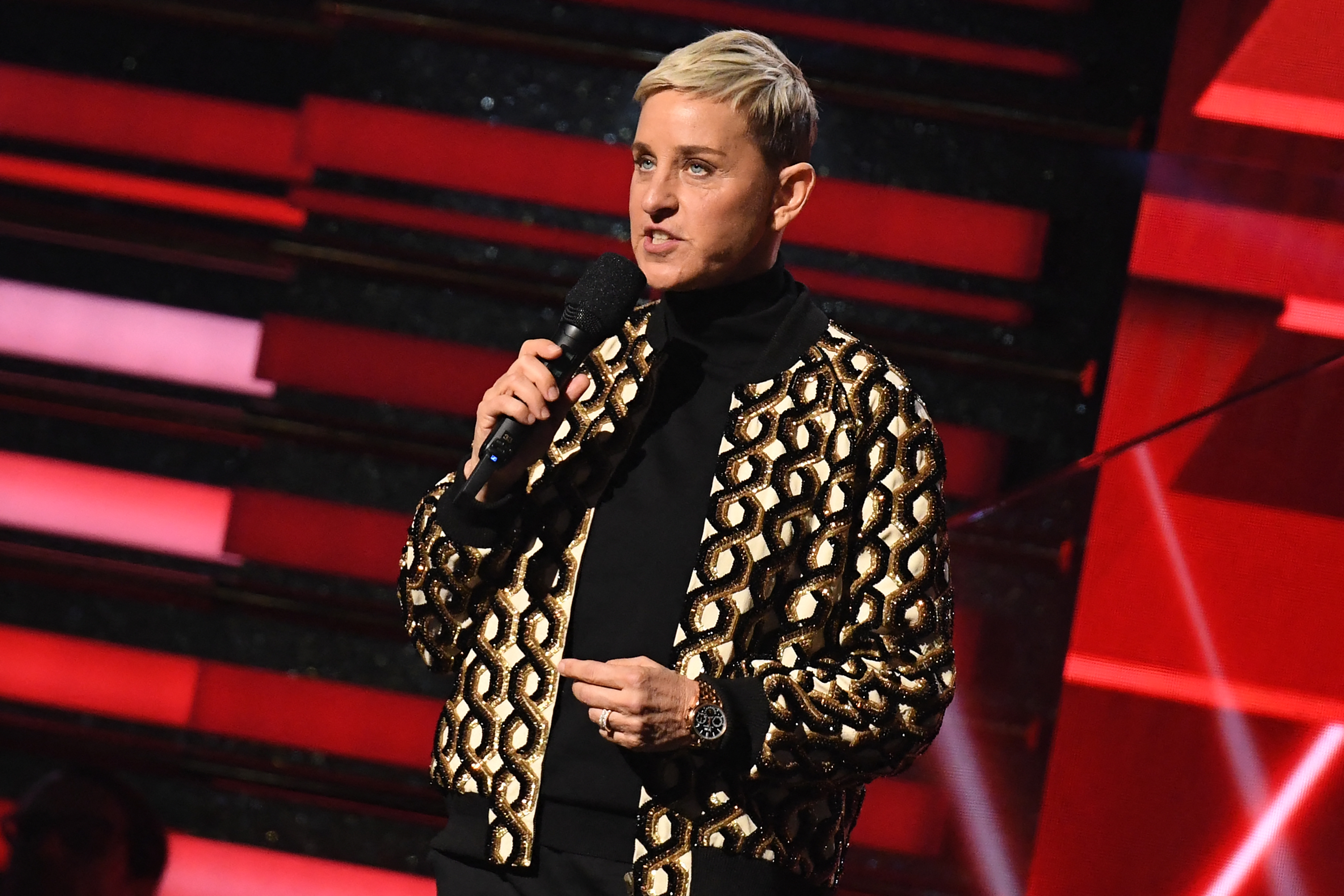 Ellen Degeneres in a stylish patterned jacket speaks on stage, holding a microphone against a backdrop of horizontal stripes