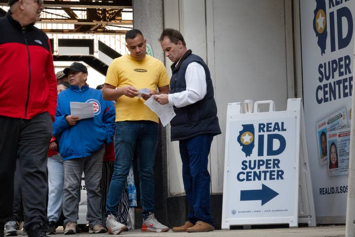 People standing in line outside a Real ID Super Center, holding documents, near a sign pointing to the center