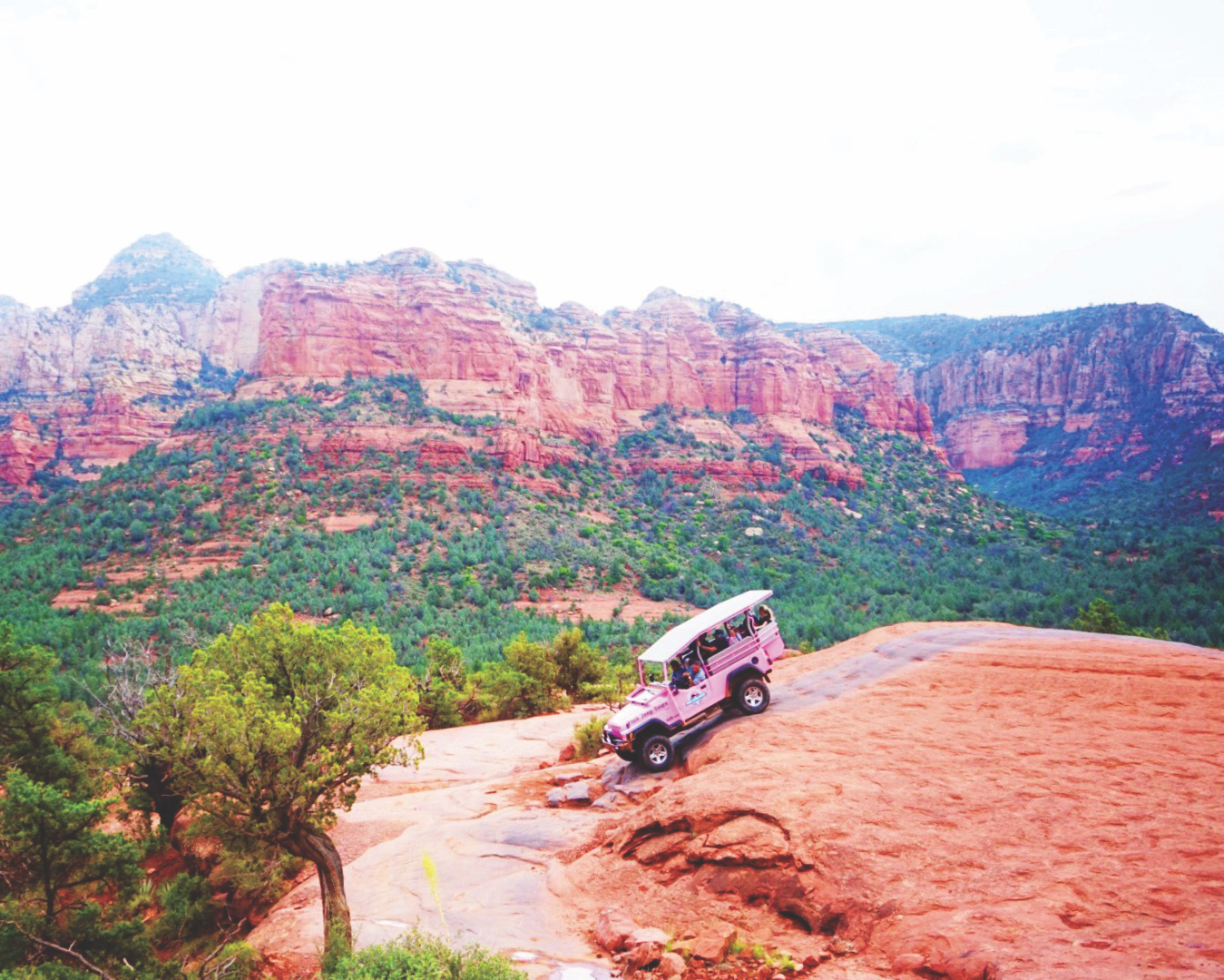 A pink Jeep is parked on a rocky ledge overlooking a vast scenic landscape with red rock formations and green vegetation
