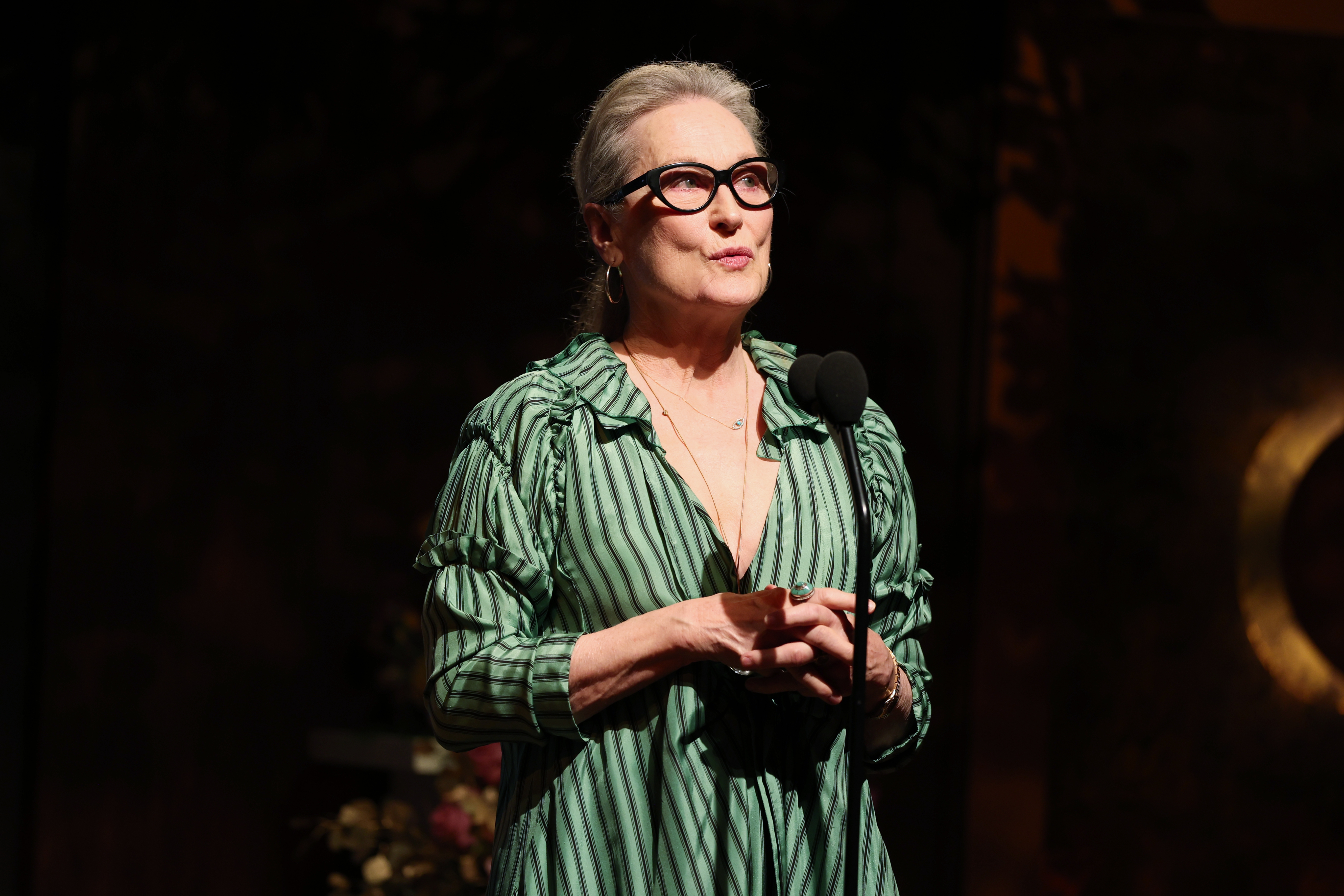 Meryl Streep stands on stage with a microphone, wearing a striped, flowing dress and glasses