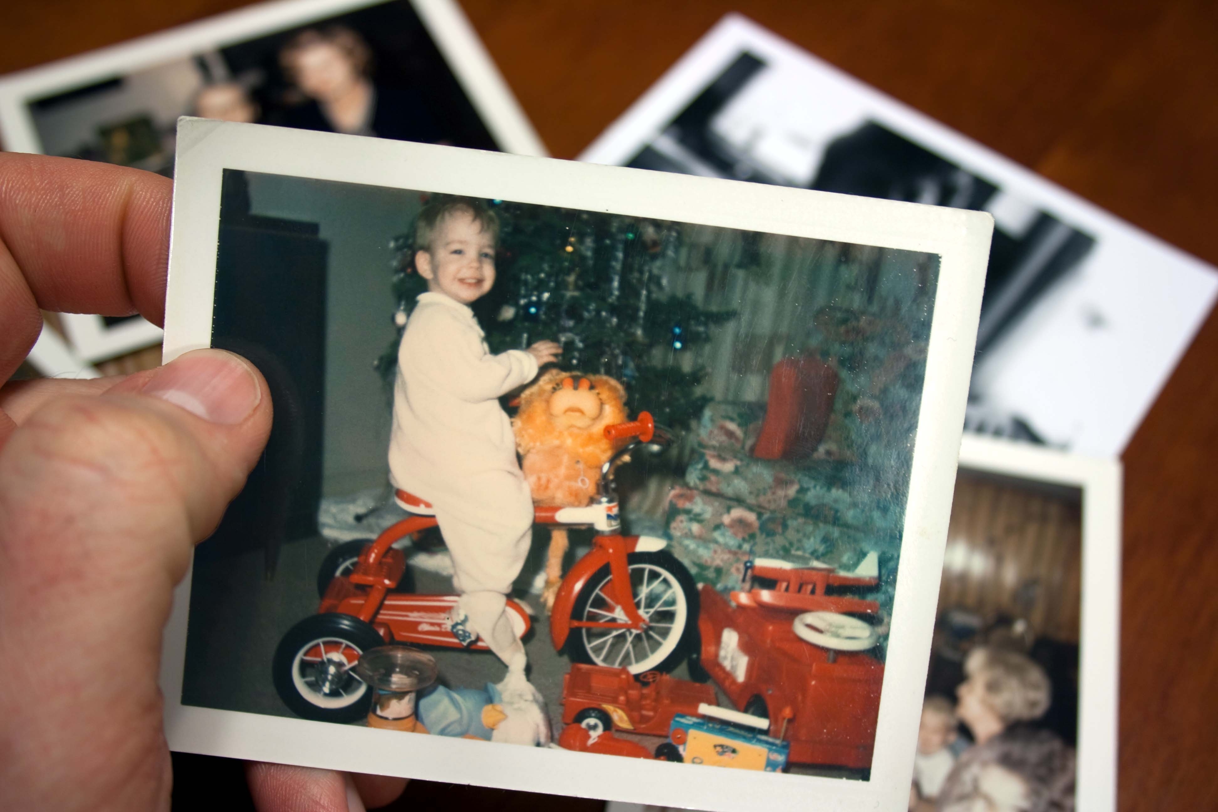 A person holds a photo of a smiling child in pajamas sitting on a tricycle next to a Christmas tree, with more photos in the background