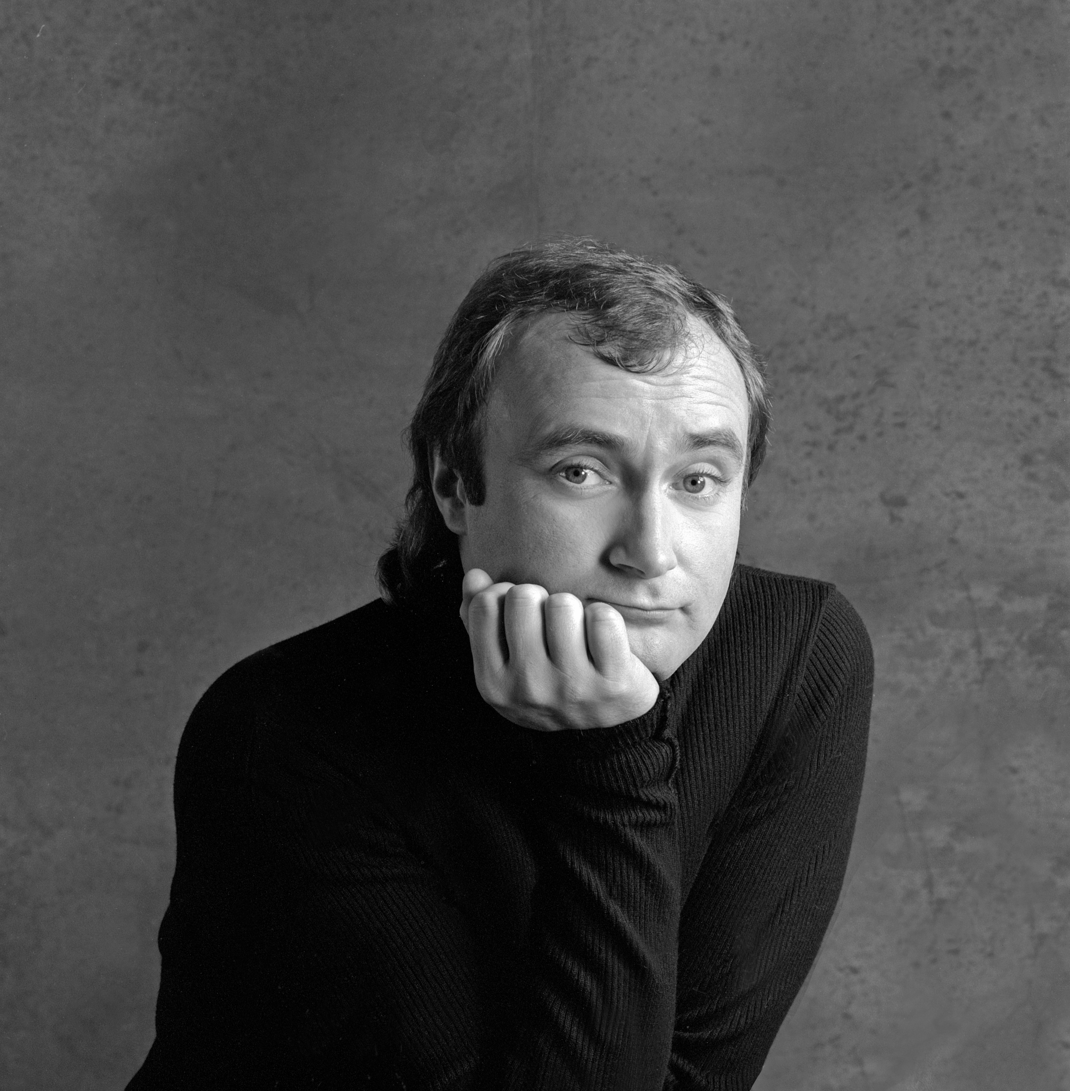 Phil Collins with a thoughtful expression, leaning on his hand, wearing a textured sweater, in a studio setting