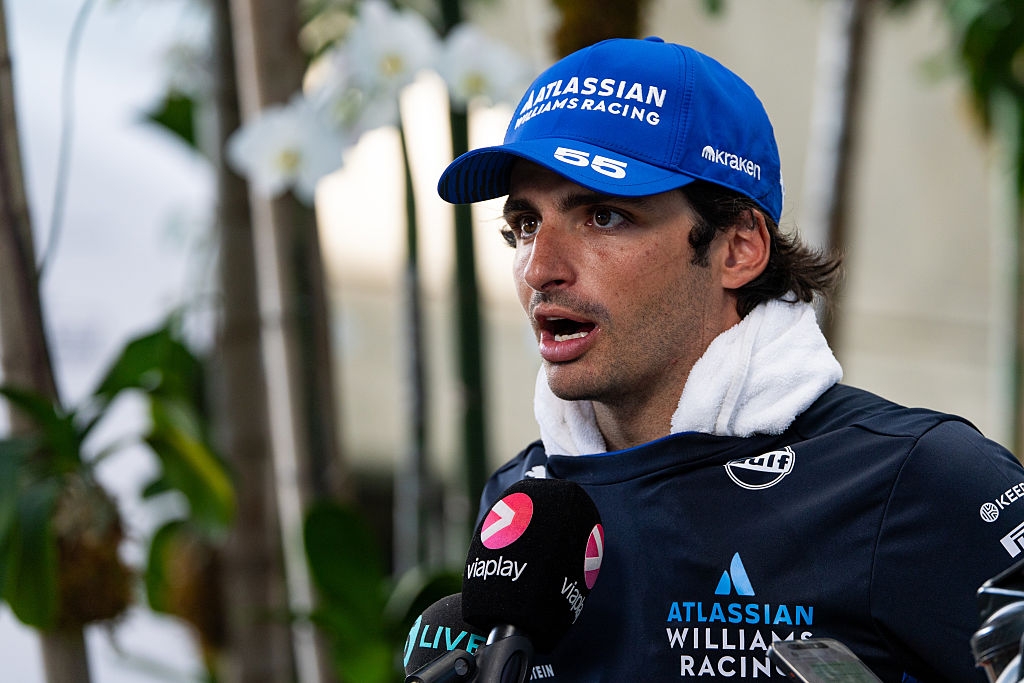 Person in a racing team jacket and cap, holding a towel, speaks to microphones in a tropical location
