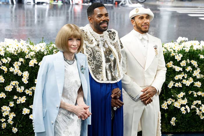 Three individuals stand on a floral-themed event carpet. One wears a pastel suit, one has ornate clothing, and one is in an elegant white suit with a beret