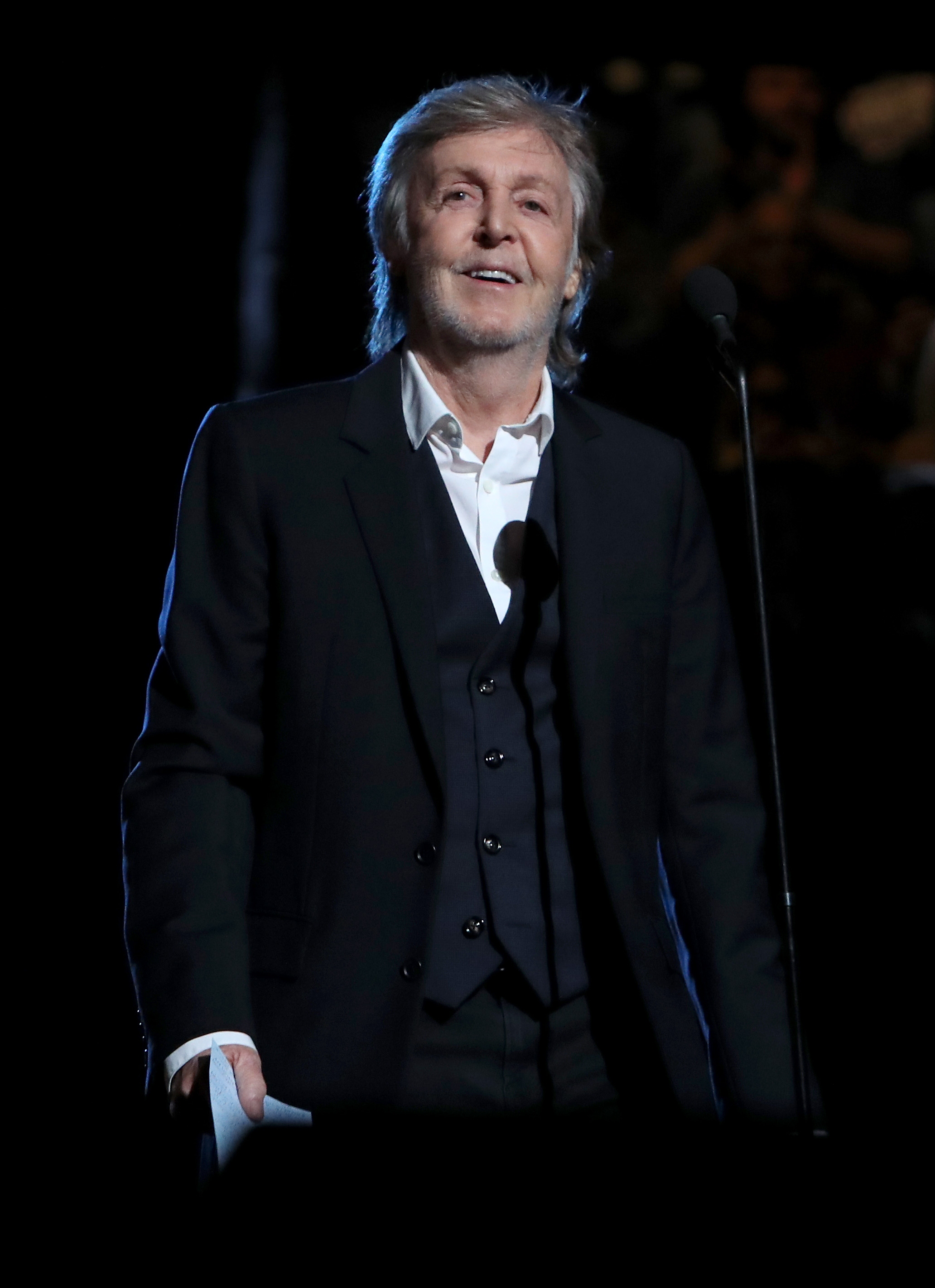 Person in a black suit and white shirt speaks at a microphone on stage