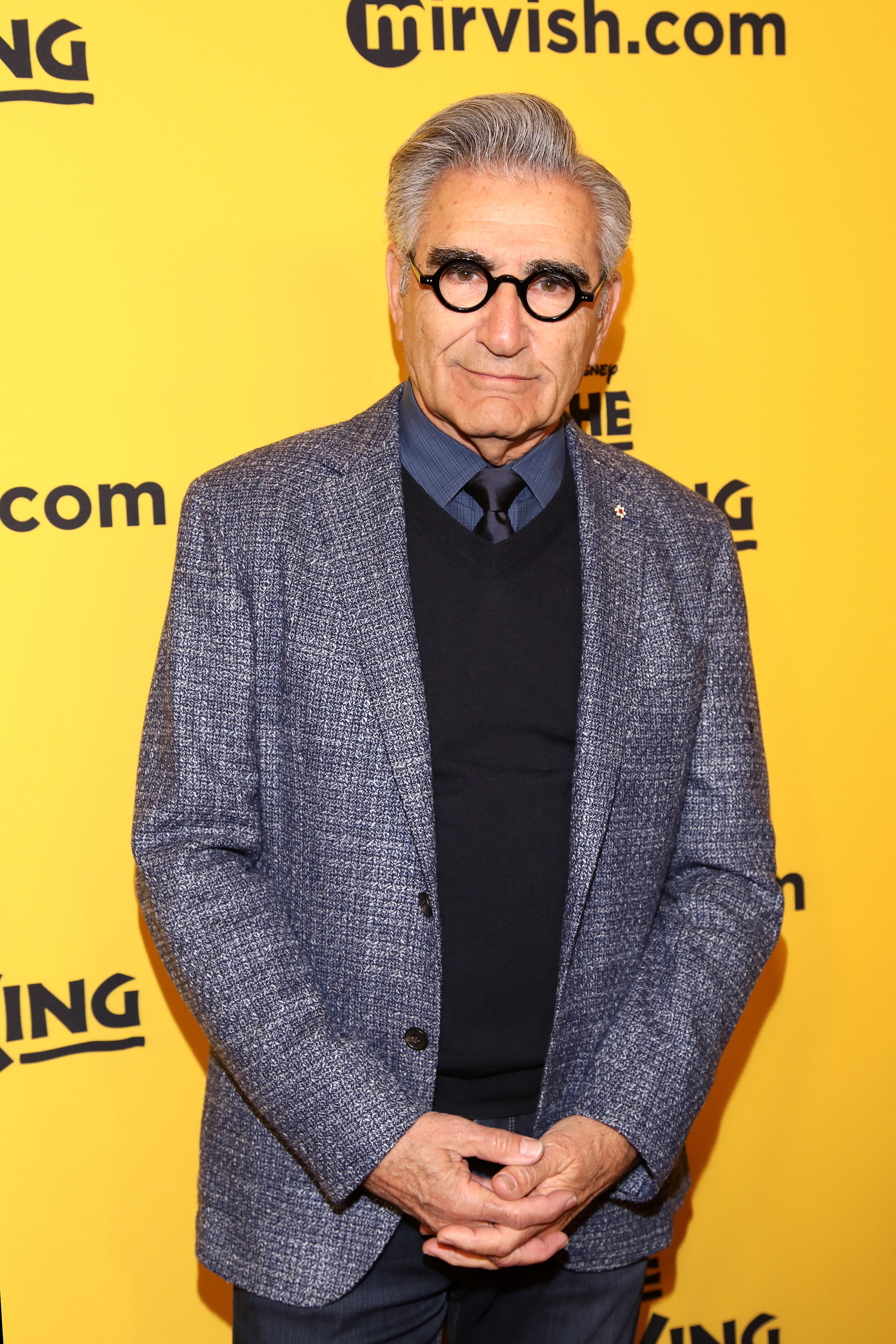 A man in a textured blazer and black sweater stands on a yellow carpet at an event, wearing round glasses, with a calm expression