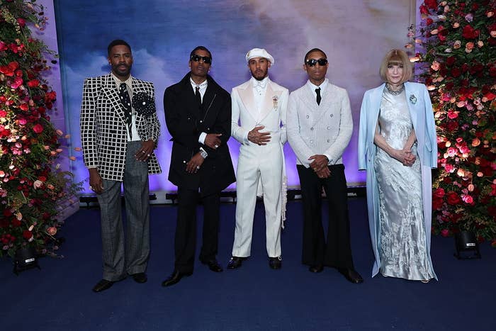 Five people pose in elegant formal wear with floral decorations in the background