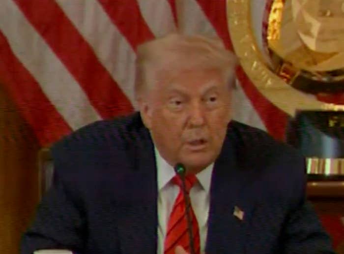 A person speaking at an event with flags in the background, dressed in a suit and tie