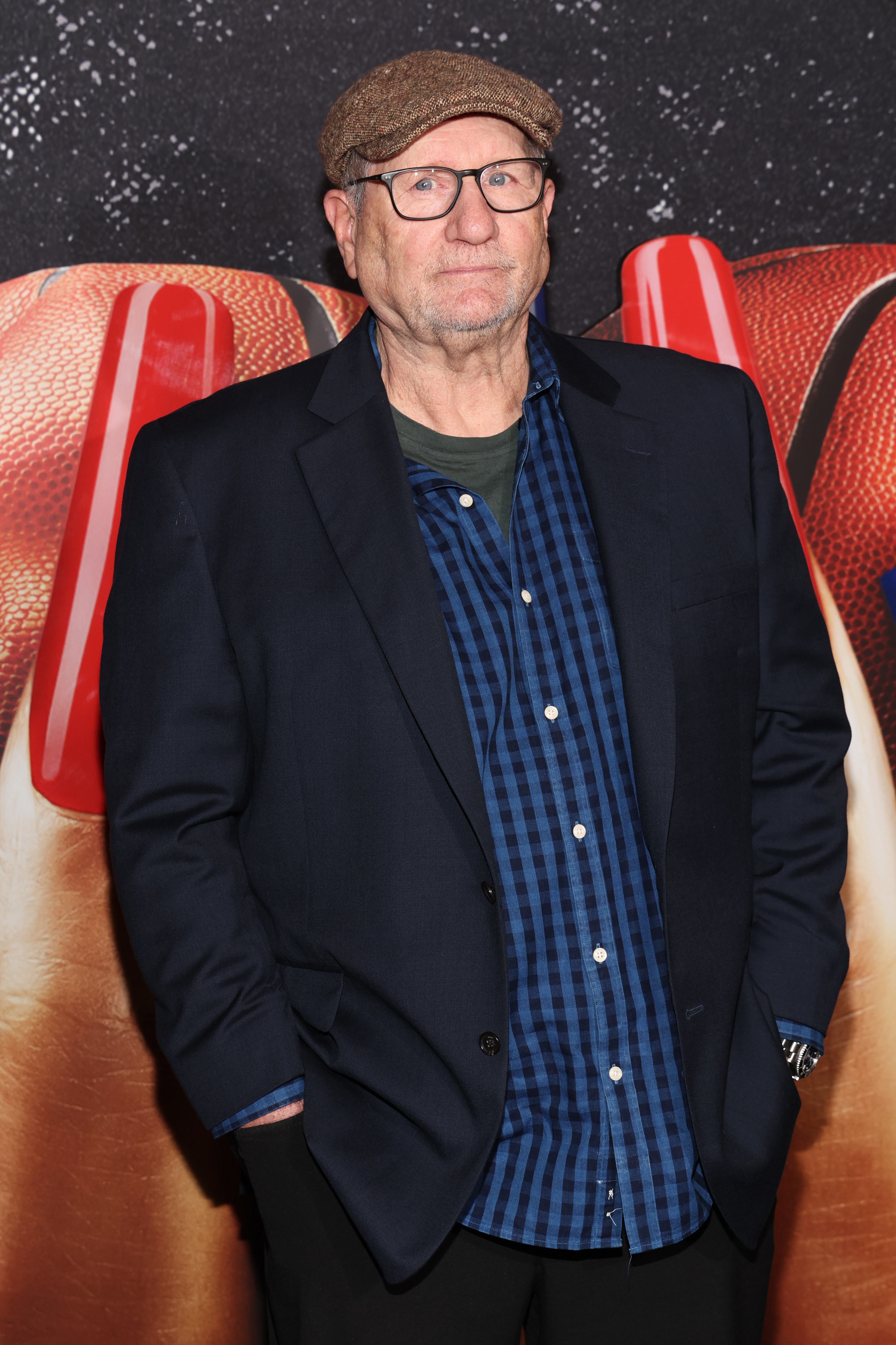 Person in flat cap and glasses wearing a blazer over a checked shirt on a red carpet