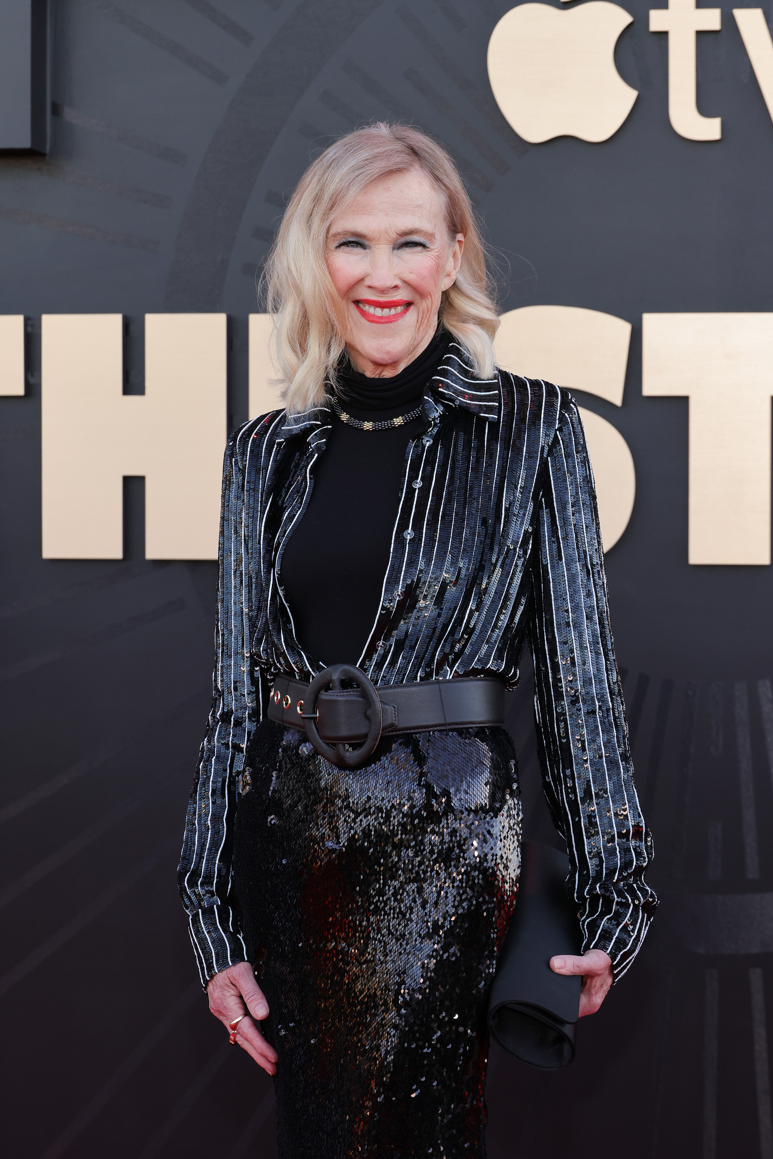 Person in sleek black sequined dress and striped jacket stands on a red carpet, smiling
