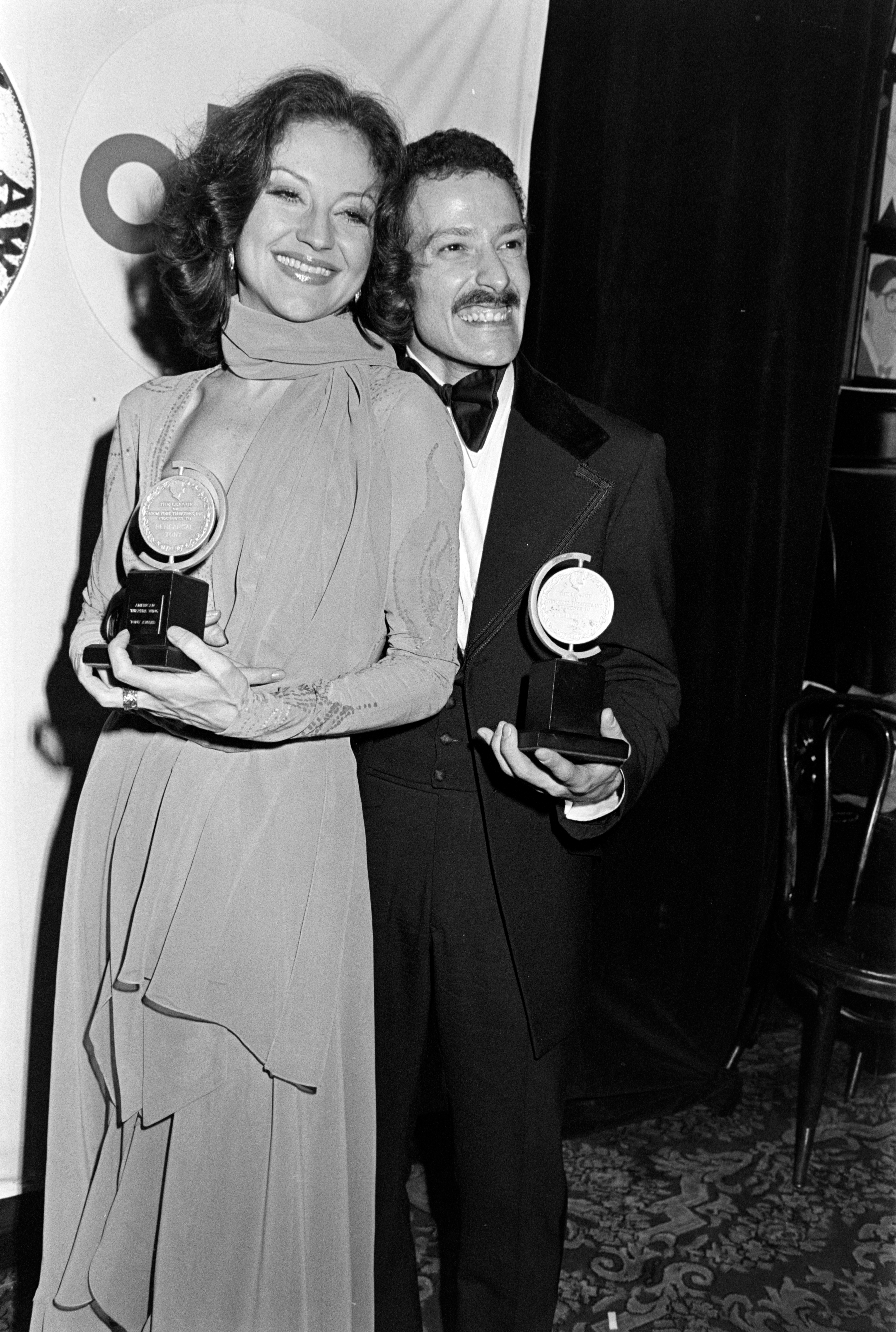Two people smiling with awards on a red carpet, woman in a draped gown, man in a tuxedo with a bow tie