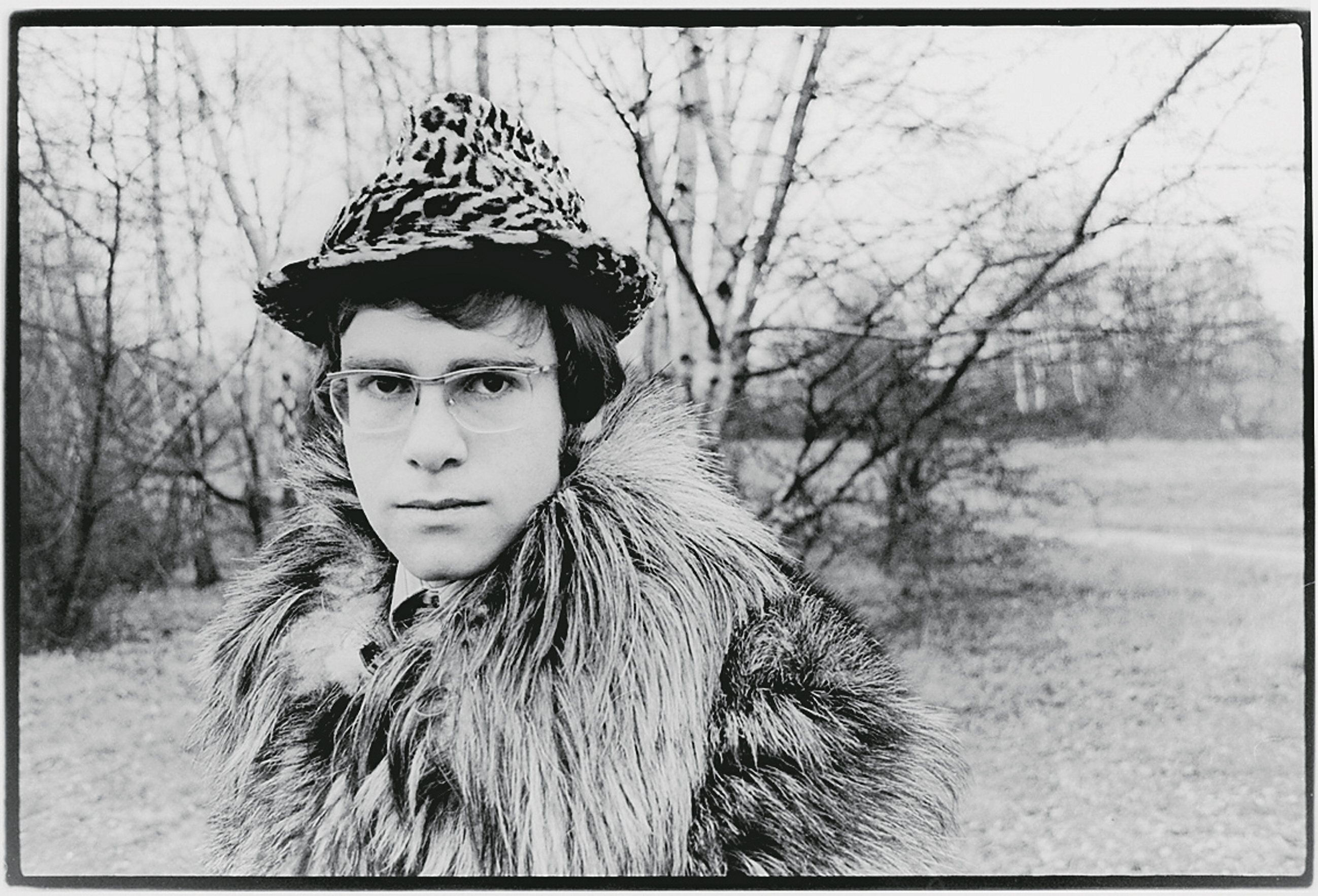 Person in glasses wearing a leopard-print hat and fur coat, standing outdoors with bare trees in the background