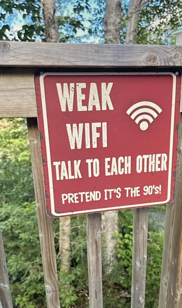 Sign on a wooden railing reads, "Weak WiFi, talk to each other. Pretend it's the 90's!" in a forested area