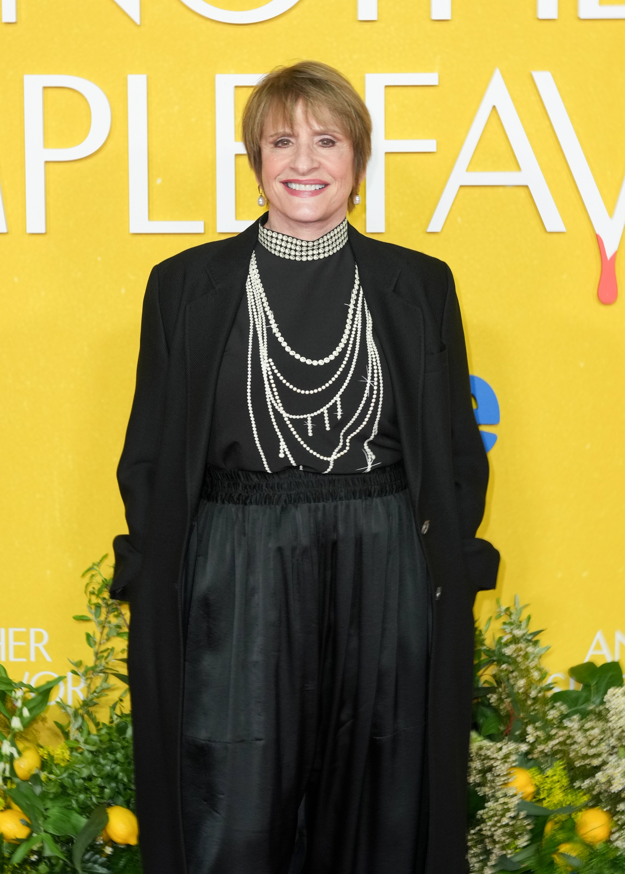 Person on red carpet in a black outfit with pearl necklaces, smiling against a yellow backdrop with floral decorations