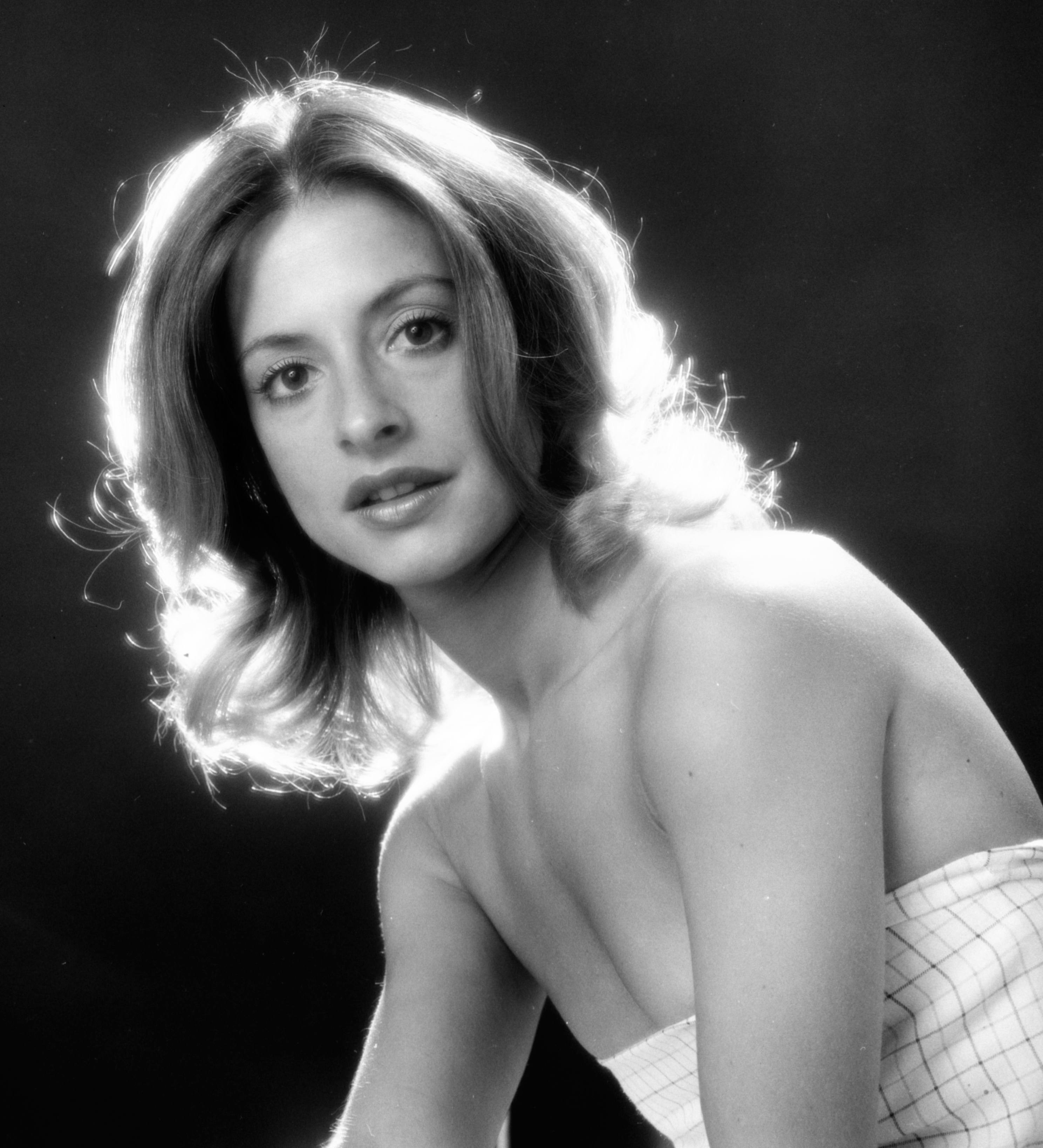 Person posing against a dark background, wearing a strapless, checkered-pattern top, with light hair styled in soft waves