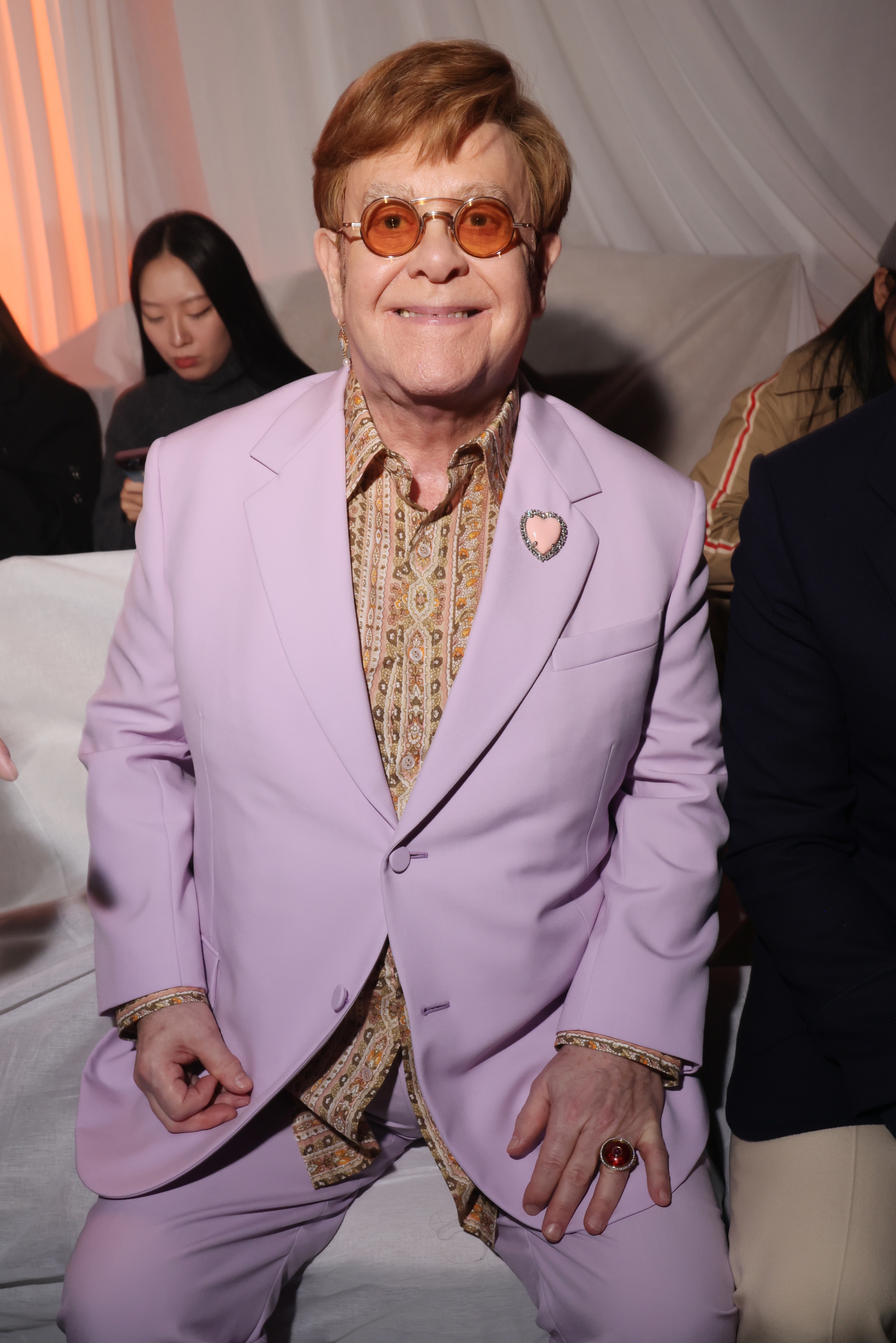 A person sits smiling, wearing a patterned shirt and stylish suit, complemented by round glasses and a ring. People are in the background