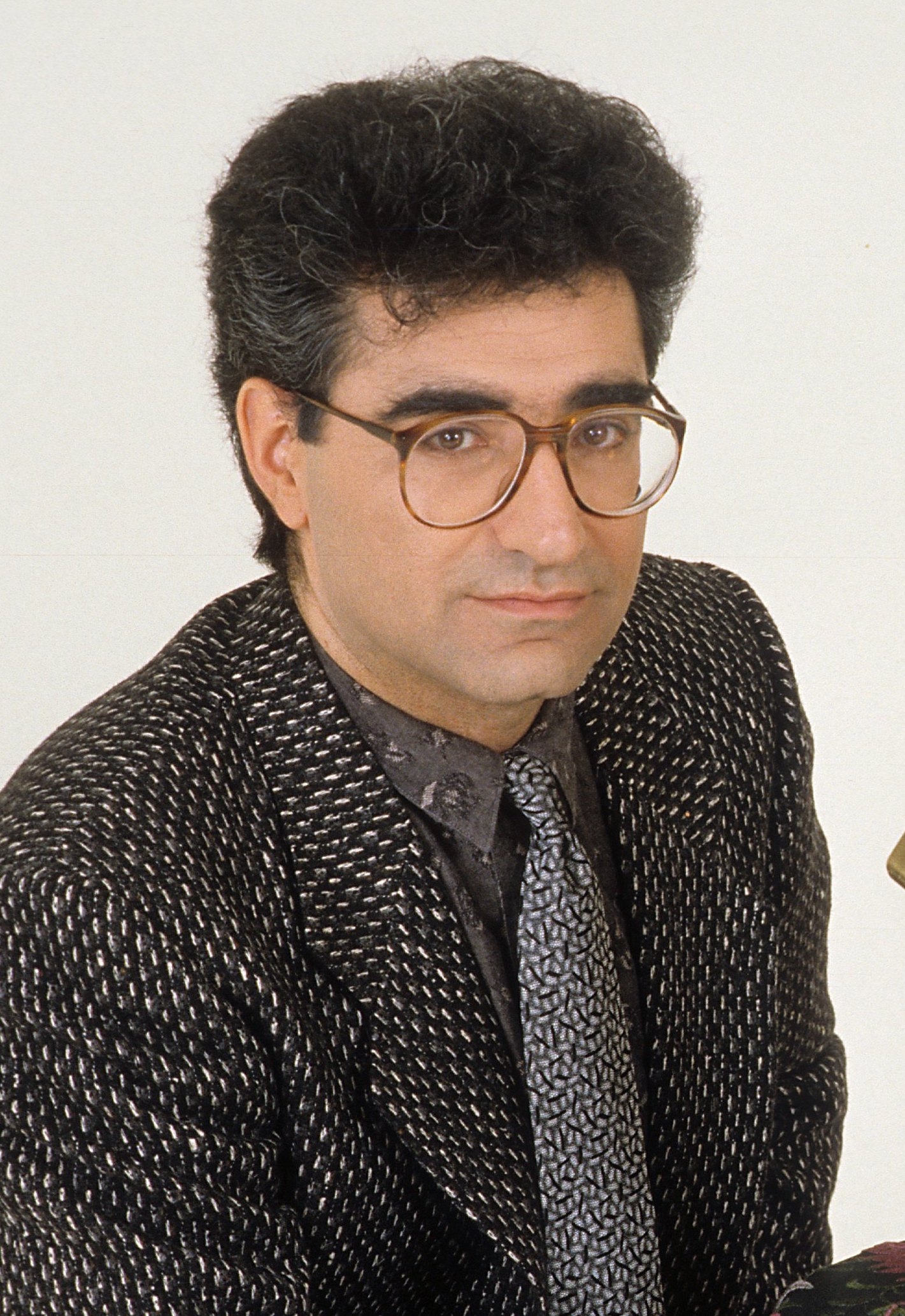 Person with curly hair, glasses, and a patterned suit and tie poses for a portrait, looking at the camera