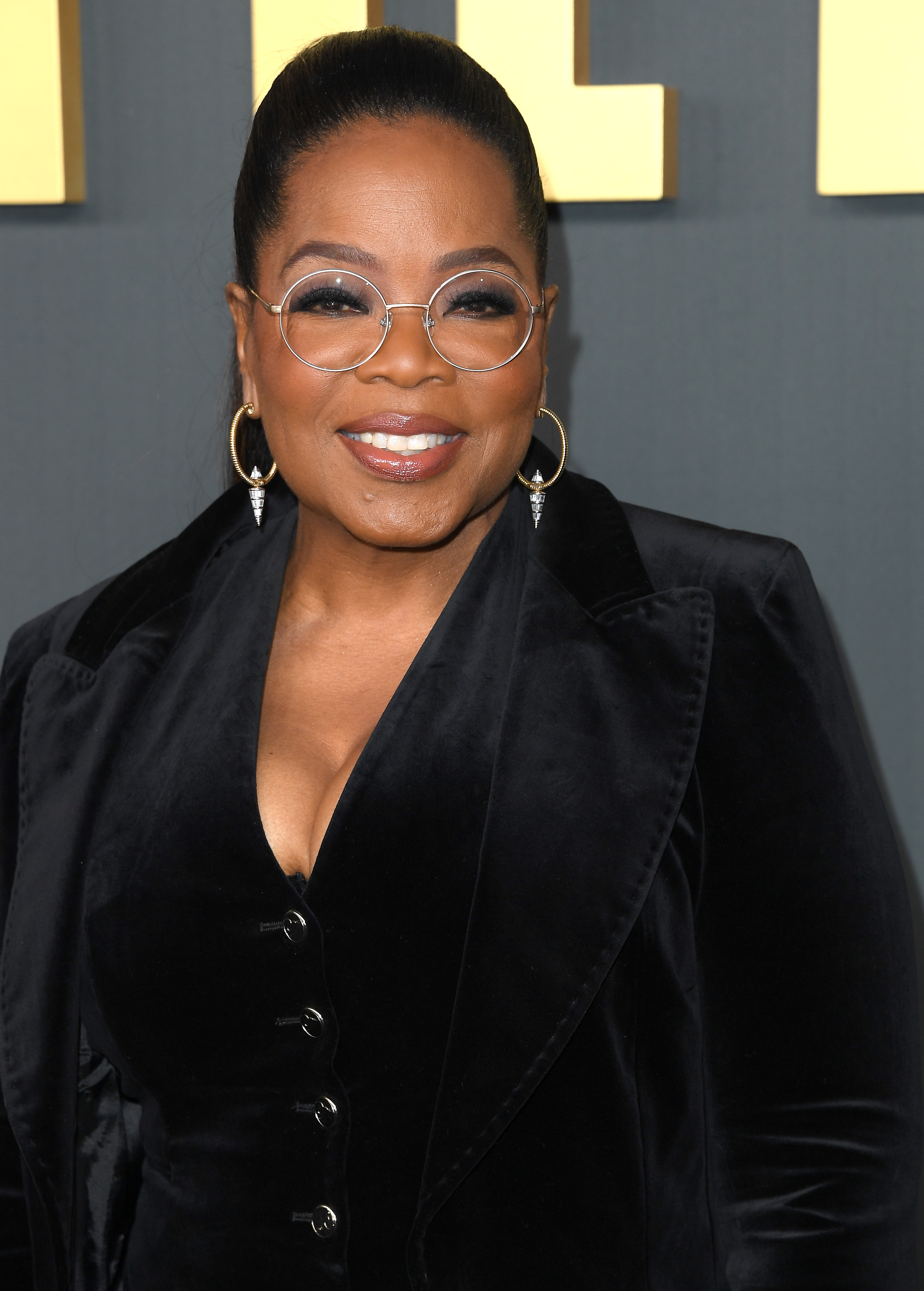 A person on a red carpet wearing a stylish velvet jacket, large hoop earrings, and round glasses, smiling for the camera