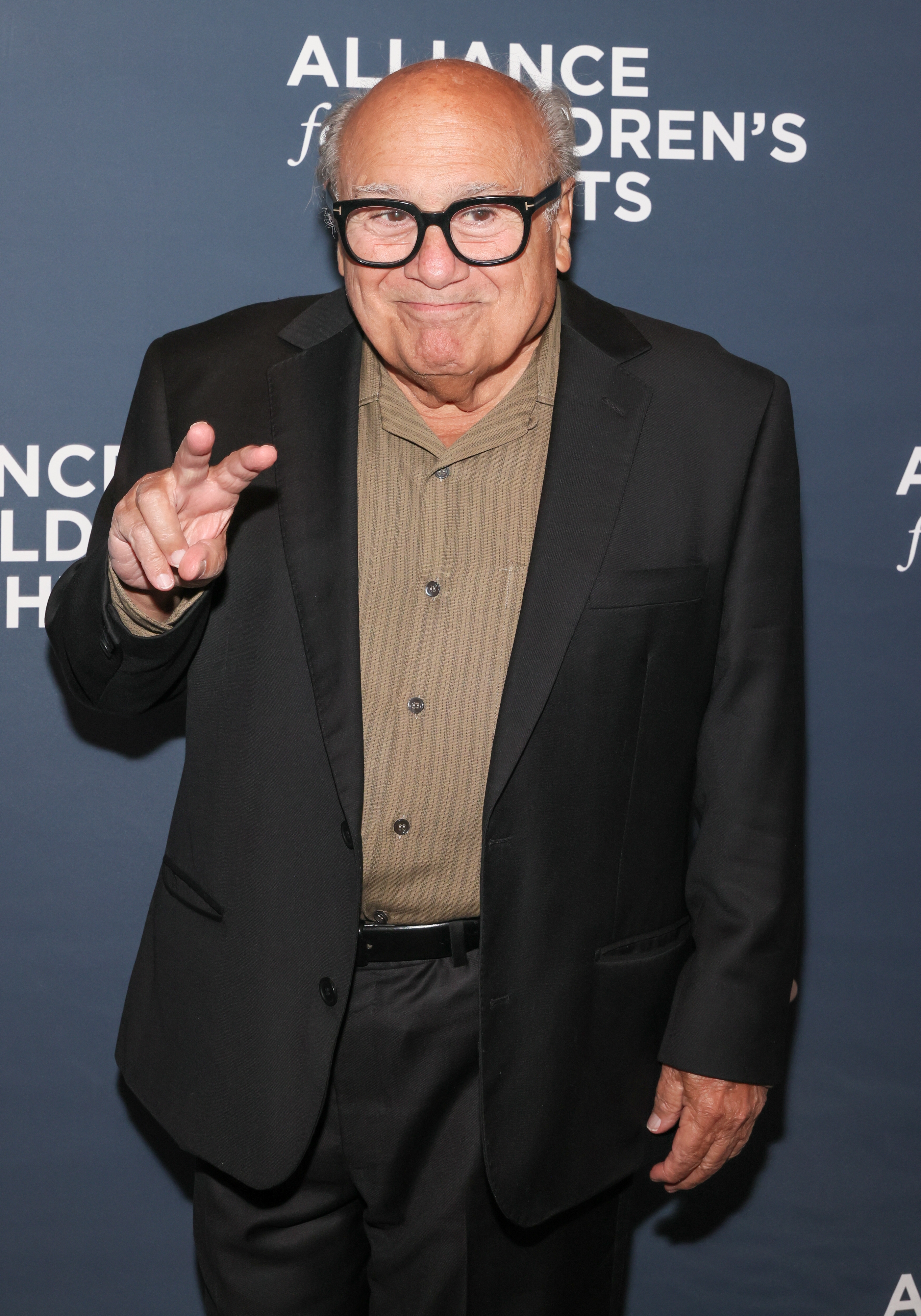 Person in a dark suit with a striped shirt and glasses poses playfully with a peace sign at a children's charity event backdrop