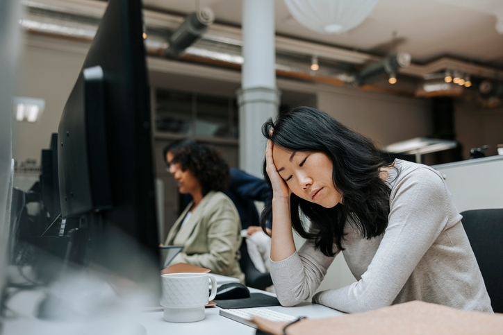 Person at a desk with head on hand, appearing tired. Computers and colleagues seen in the background, suggesting an office setting