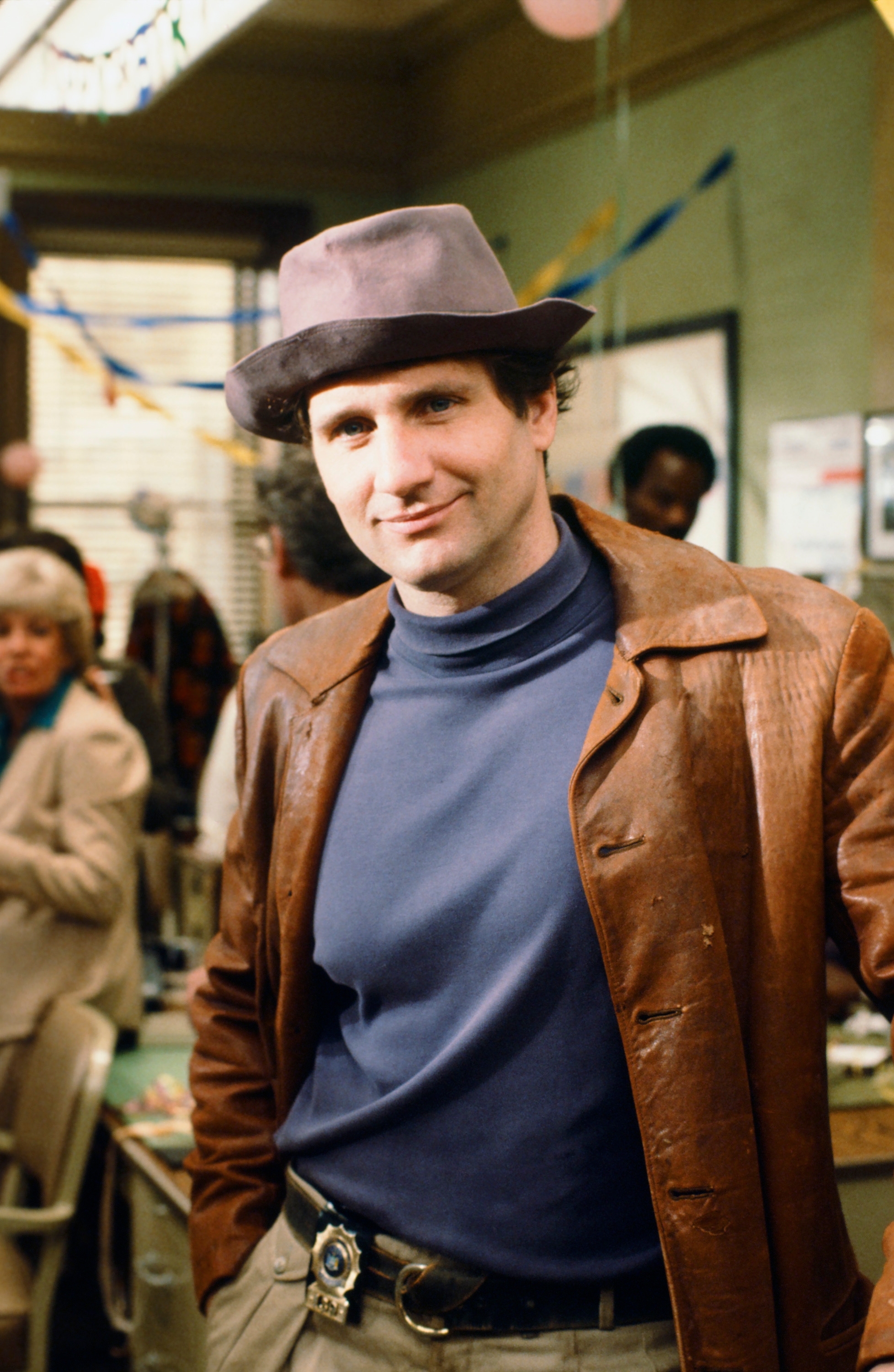 A person in a 1970s-style ensemble with a brown leather coat and fedora, standing confidently at a lively indoor gathering