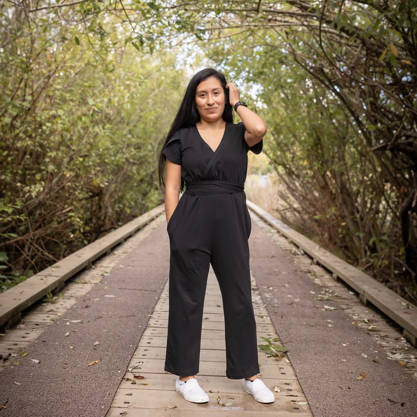 Person stands on a wooden path surrounded by trees, wearing a stylish black jumpsuit and white sneakers, looking confidently at the camera