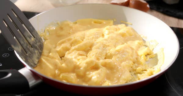 Scrambled eggs cooking in a frying pan, partially stirred with a spatula, with melted cheese on top