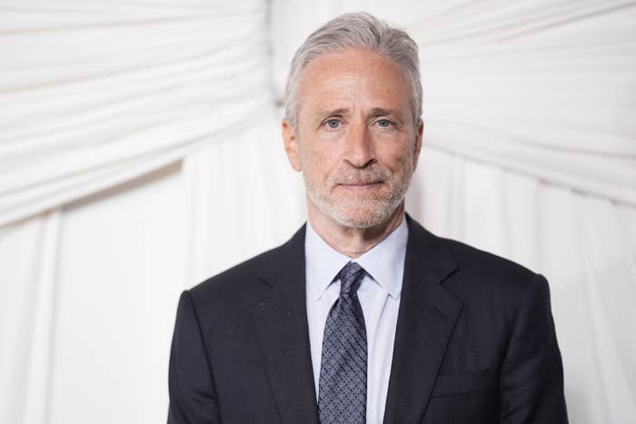 A person in a suit with a neutral expression stands against a draped background