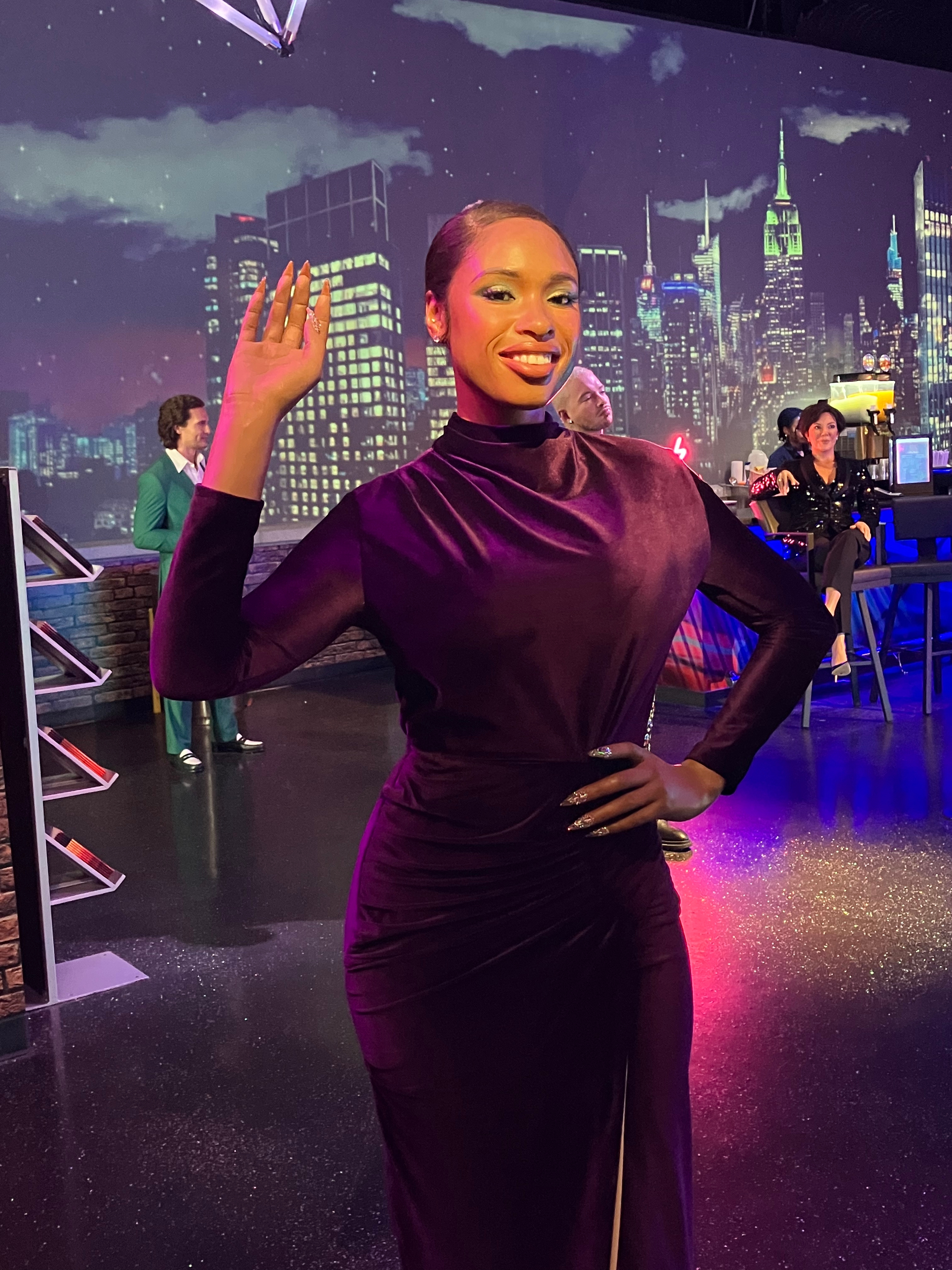 Person in a sleek, long-sleeved gown poses with a city skyline backdrop, waving and smiling