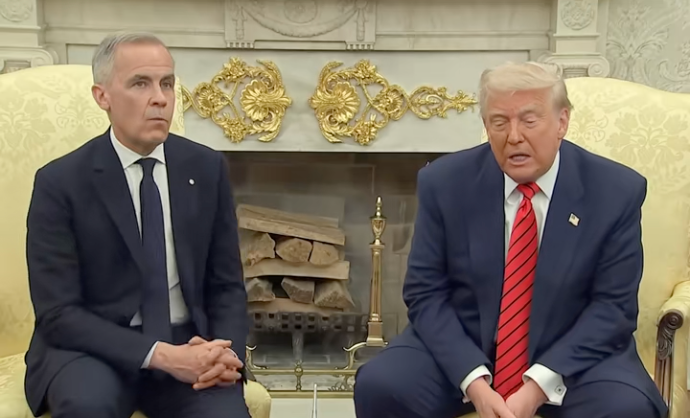 Two men sit on a couch in a formal setting. One is wearing a suit and tie; the other is in a suit with a striped tie. Fireplace in the background