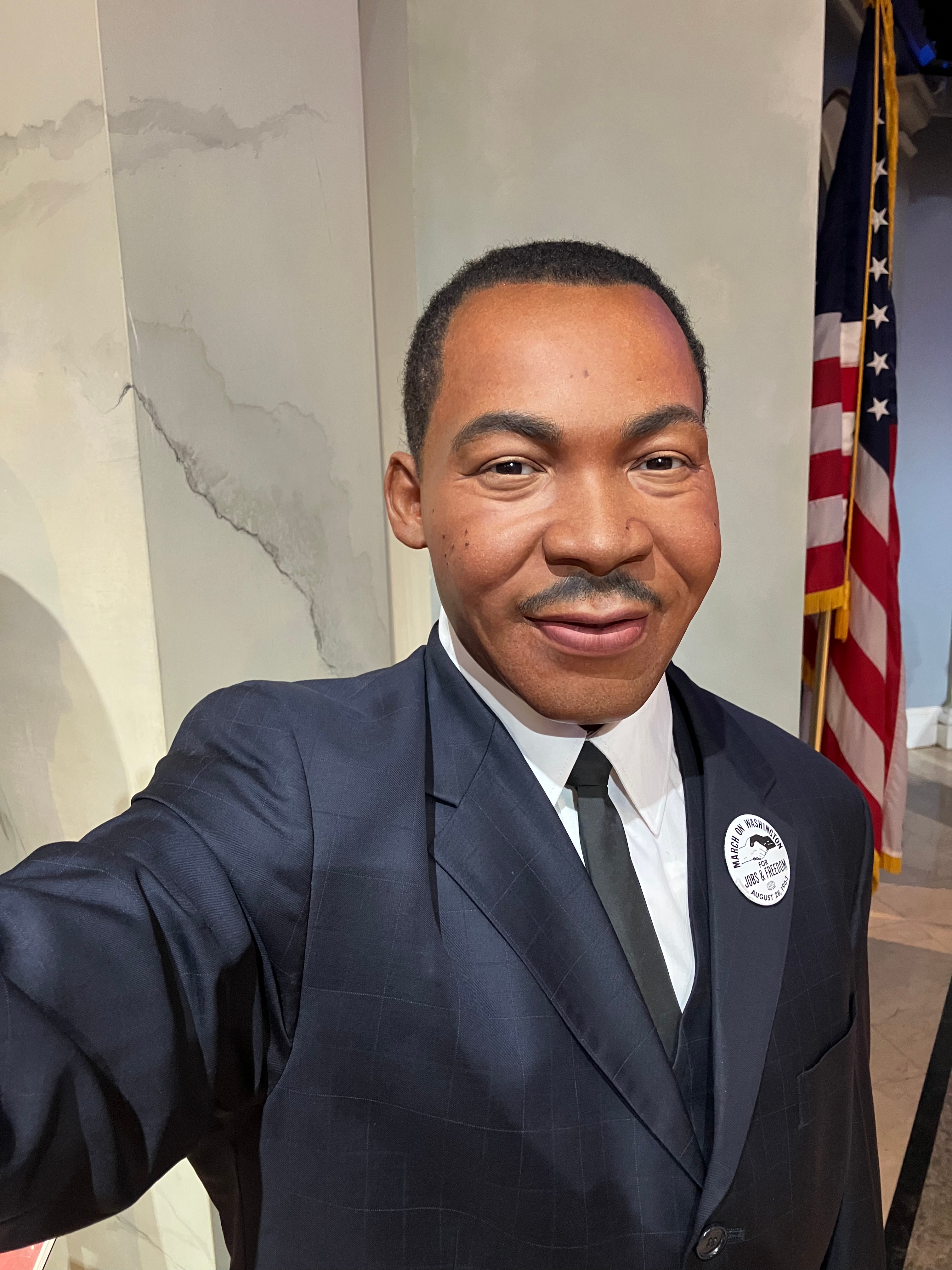 A wax figure of a well-dressed man in a suit and tie with a button on his lapel stands beside an American flag in a museum display