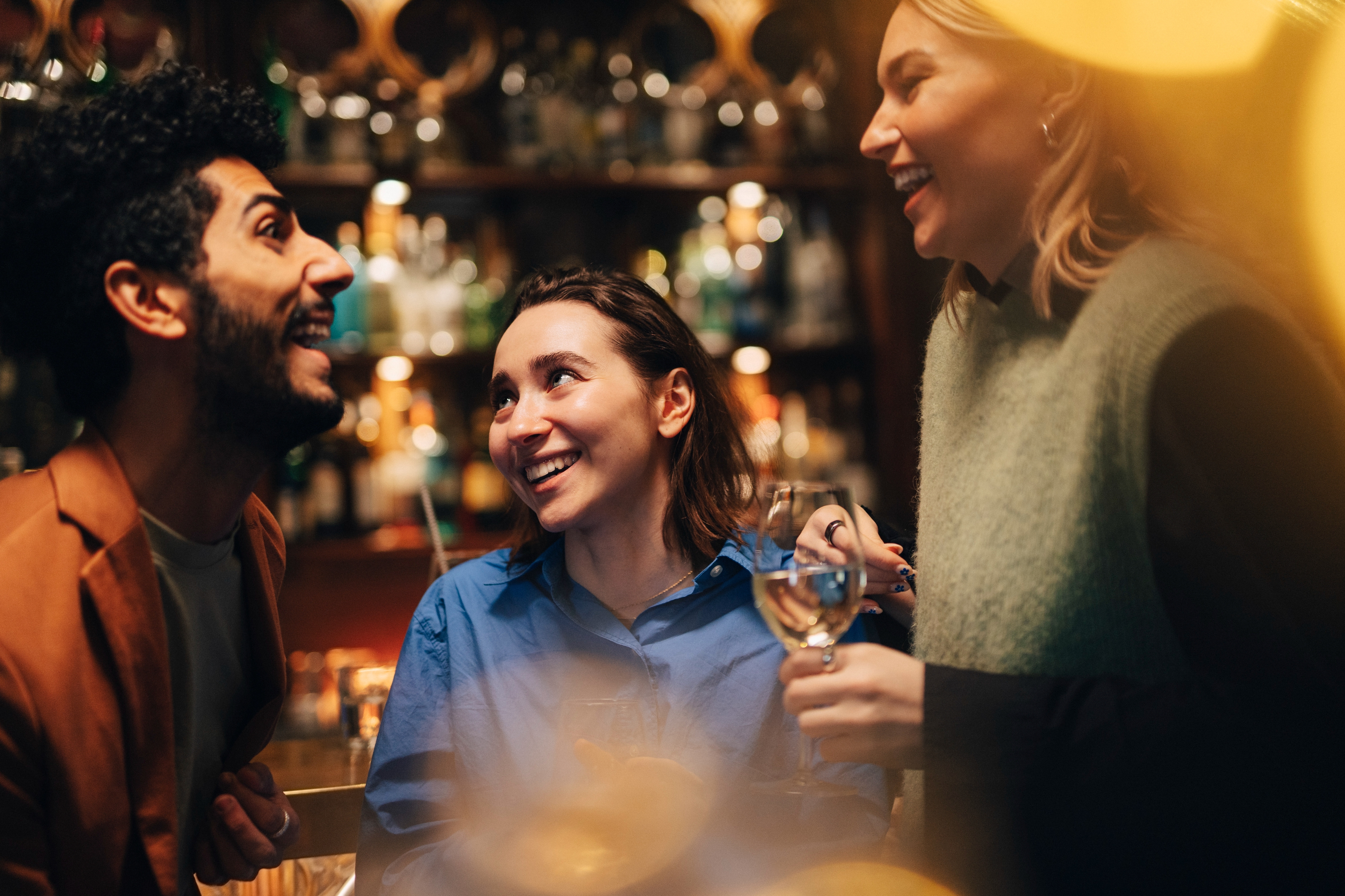 Tre personer ler og chatter på en bar, kledd i uformelle klær, med varm omgivelsesbelysning som forbedrer scenen