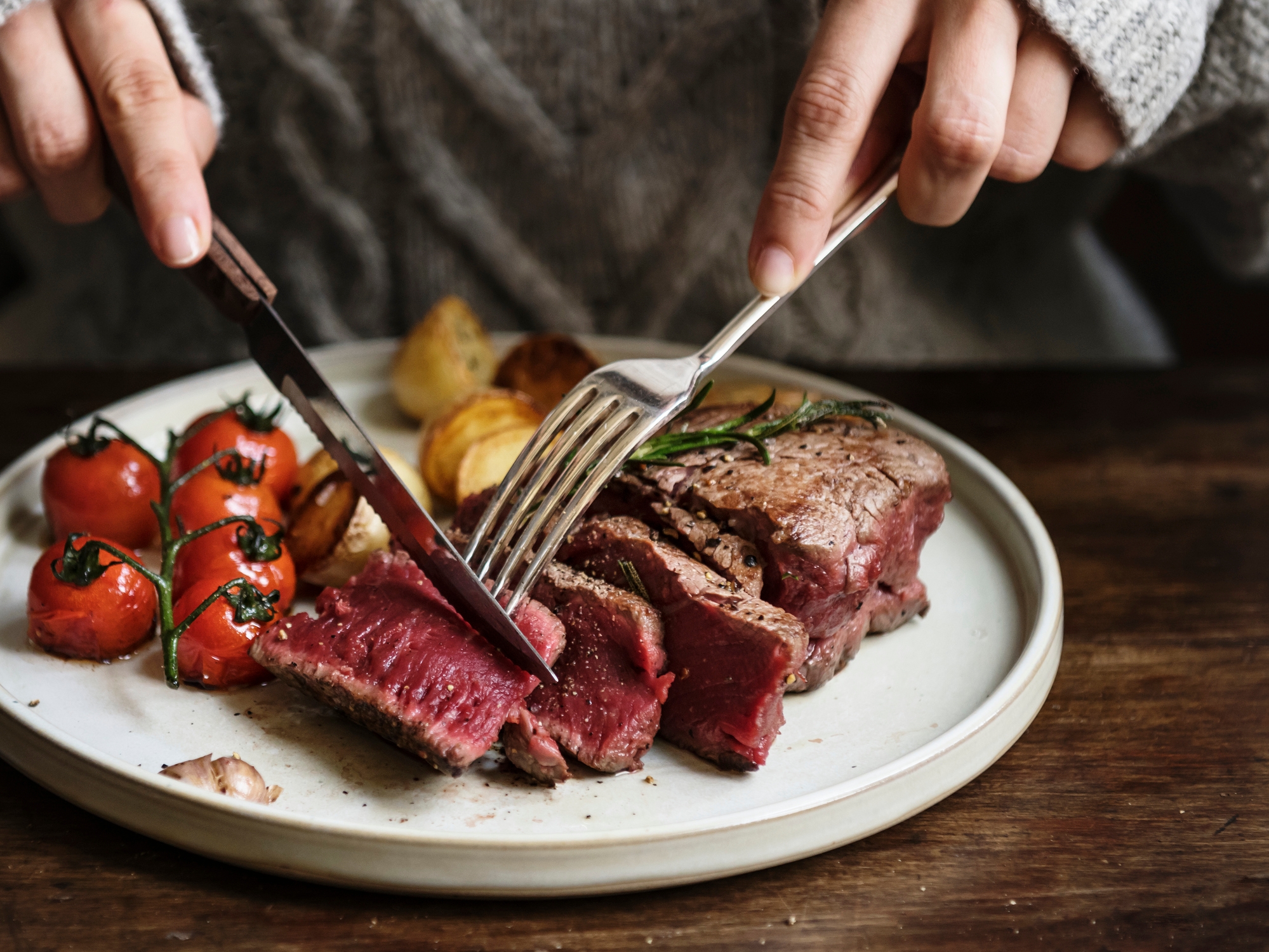 Person cutting into a medium-rare steak on a plate with roasted tomatoes and potatoes