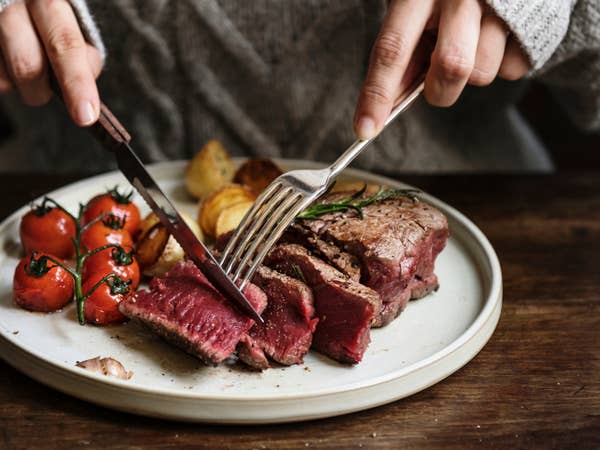 Person cutting into a medium-rare steak on a plate with roasted tomatoes and potatoes