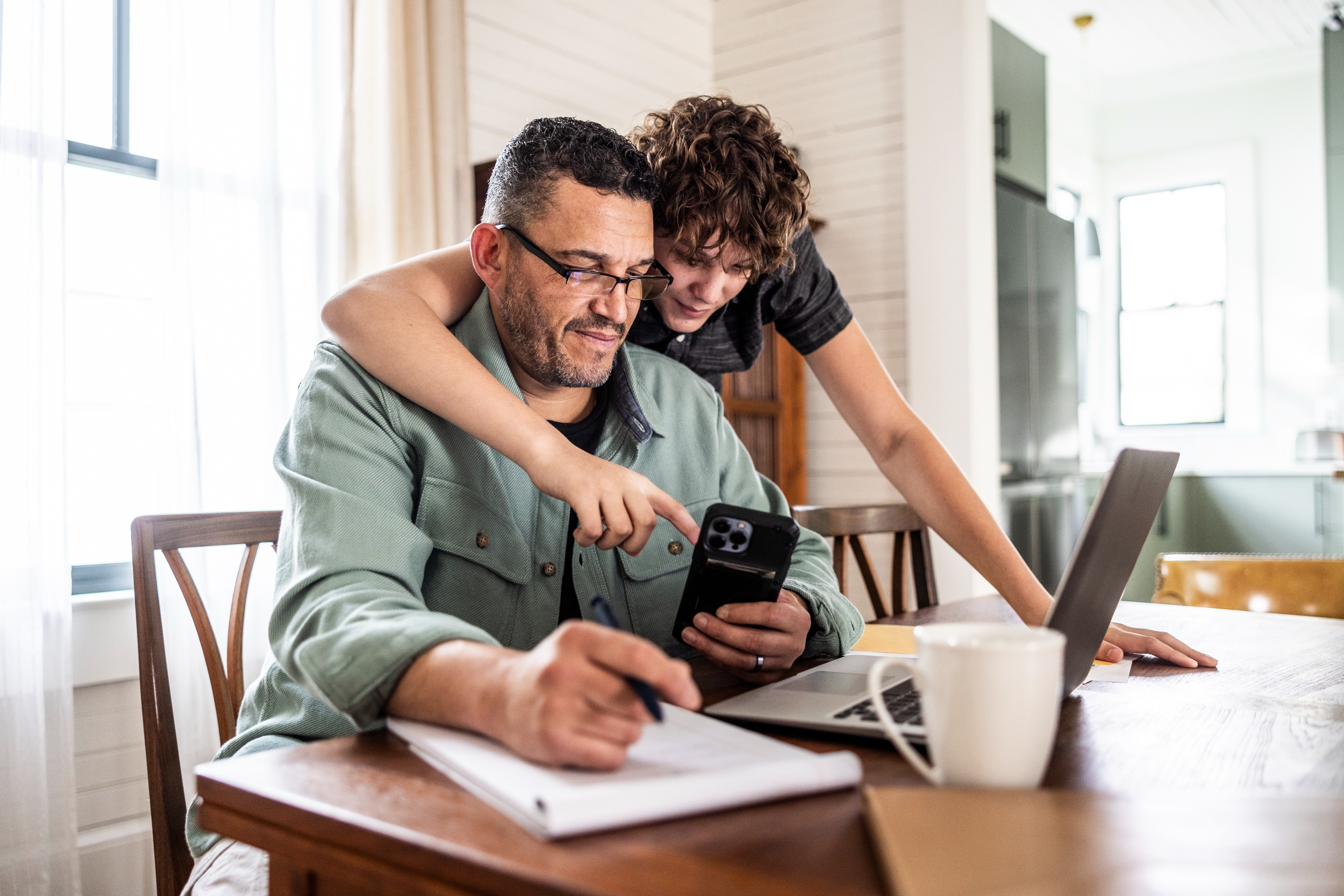 Smilende person som sitter ved et bord med en bærbar PC og notisblokk, mens en annen person står og peker på en telefon. Begge virker avslappede og nyter øyeblikket