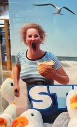 A woman poses next to a large seagull poster with her mouth open, surrounded by stuffed seagulls, humorously mimicking the seagull's expression