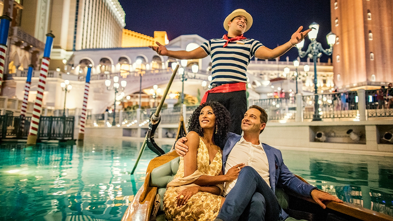 A couple sits in a gondola at a Venetian-themed resort. A gondolier stands behind them, singing passionately