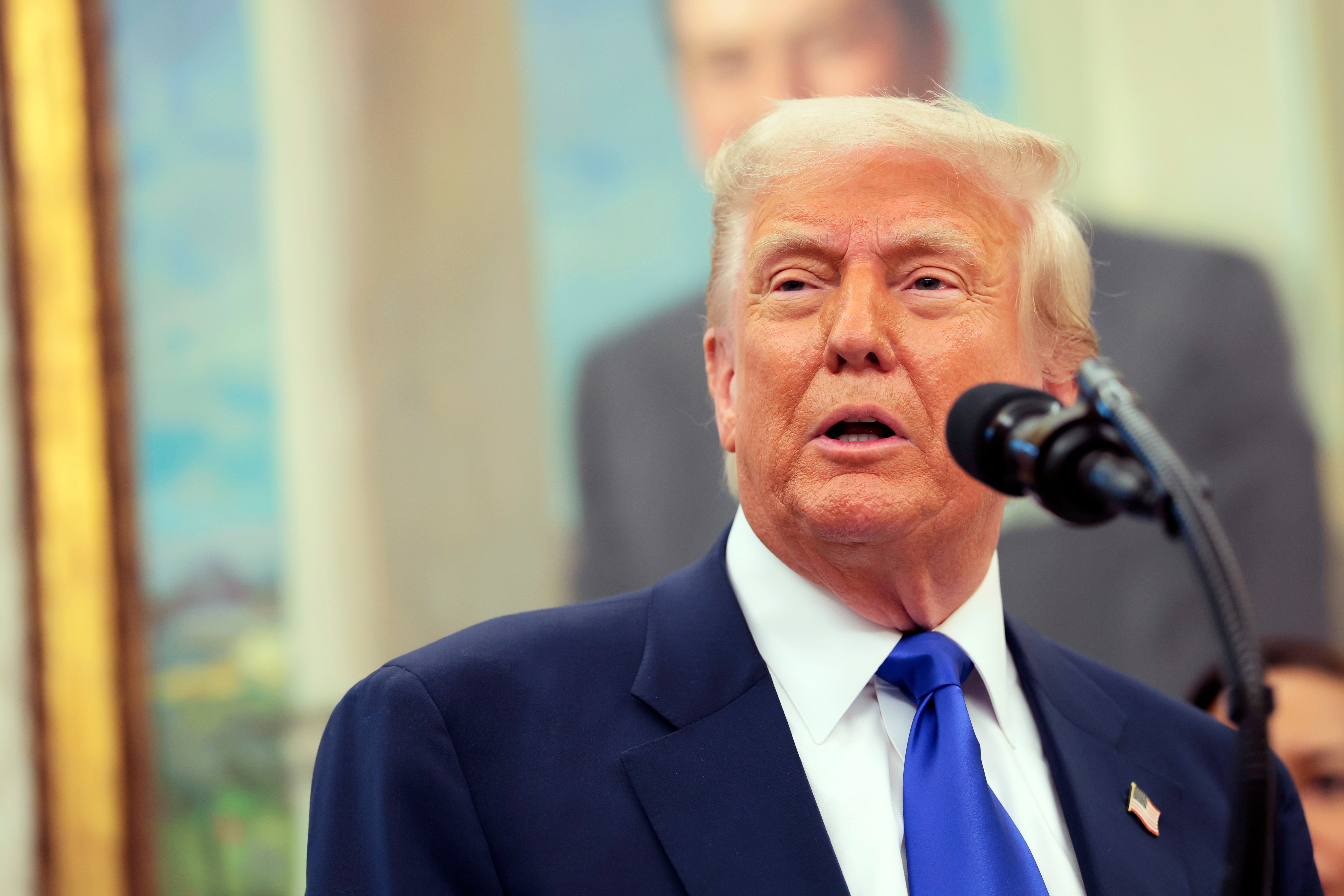 Man in a suit and tie speaks at a microphone in front of a blurred portrait background