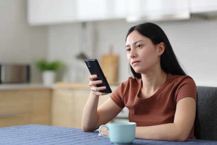 Person in a kitchen gazing at a smartphone while seated at a table with a mug in front of them