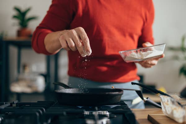 Person cooking, sprinkling flour into a pan on a stove, with a kitchen in the background