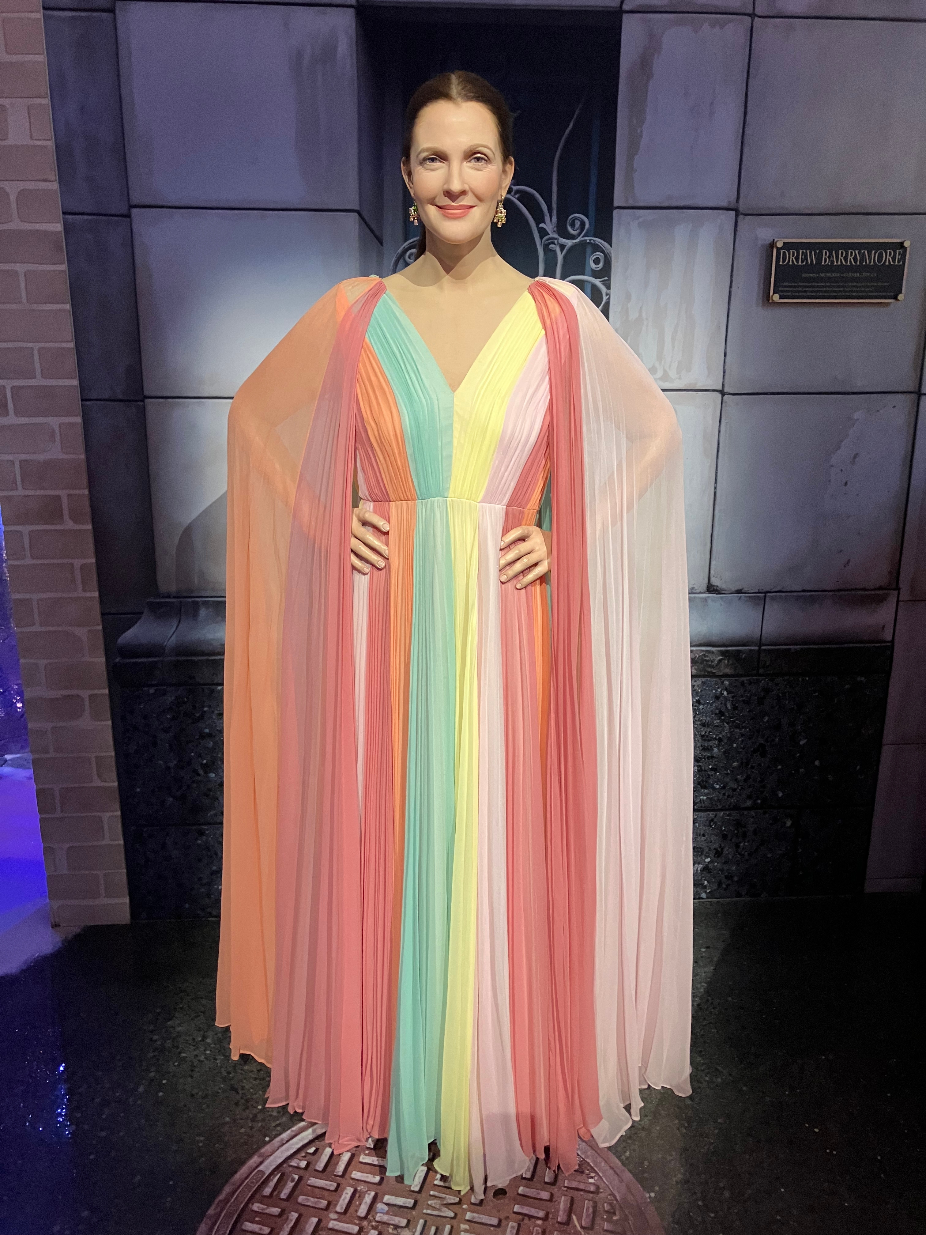 Person in a vibrant, flowing gown with draped sleeves, standing on a red carpet. They're smiling confidently with hands on hips