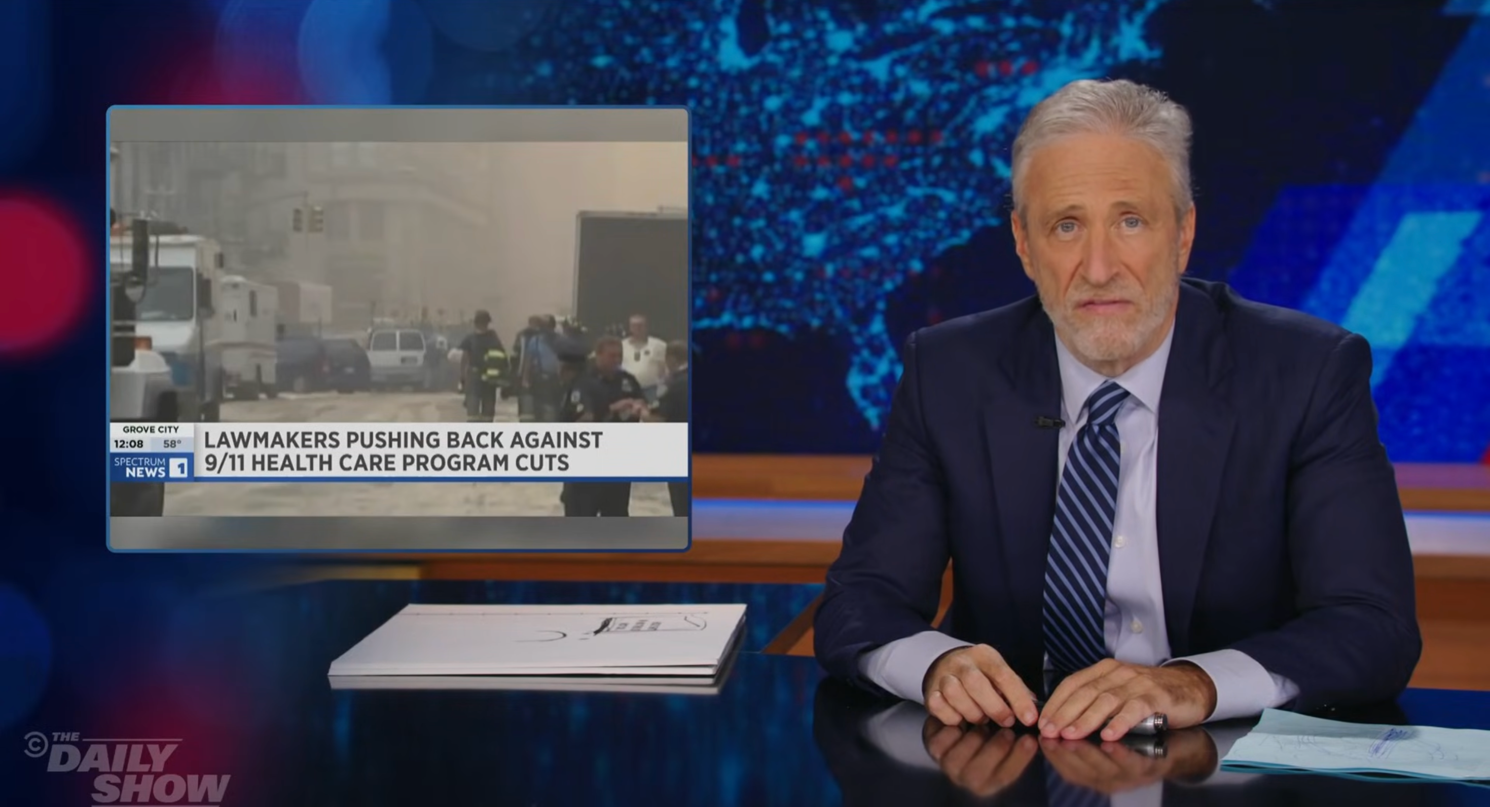 A TV host in a suit sits at a desk with a news segment on lawmakers opposing 9/11 healthcare cuts displayed on a screen beside him