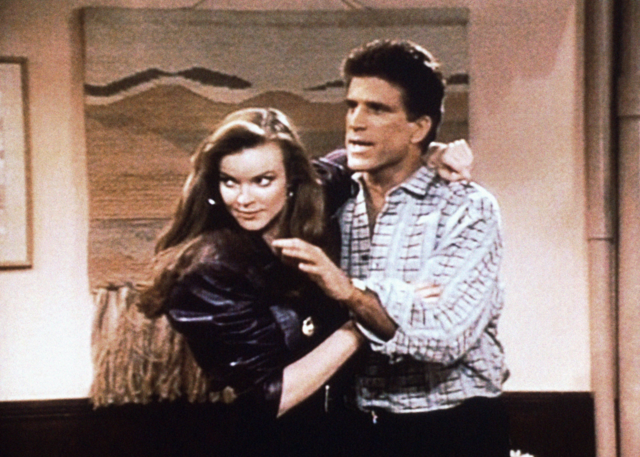 Two actors in a comedic scene; the man in a casual shirt gestures while embracing the woman in a stylish jacket